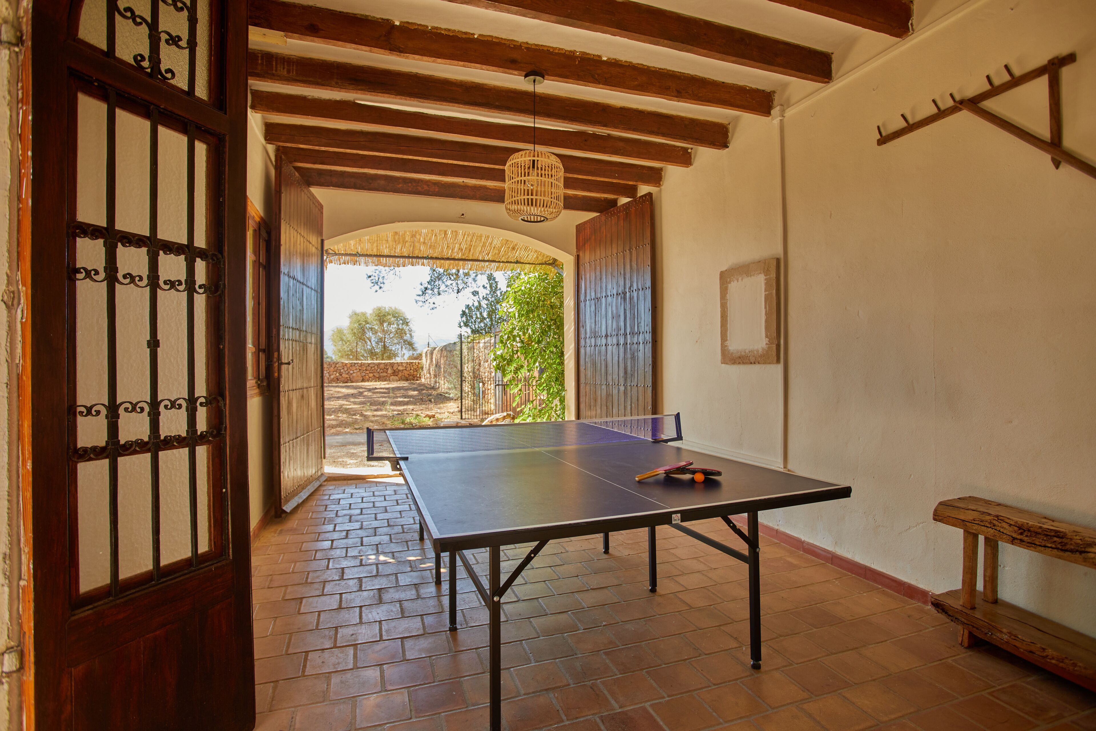A ping pong table in a room with a wooden bench, a hanging light fixture, and a view through an open door.
