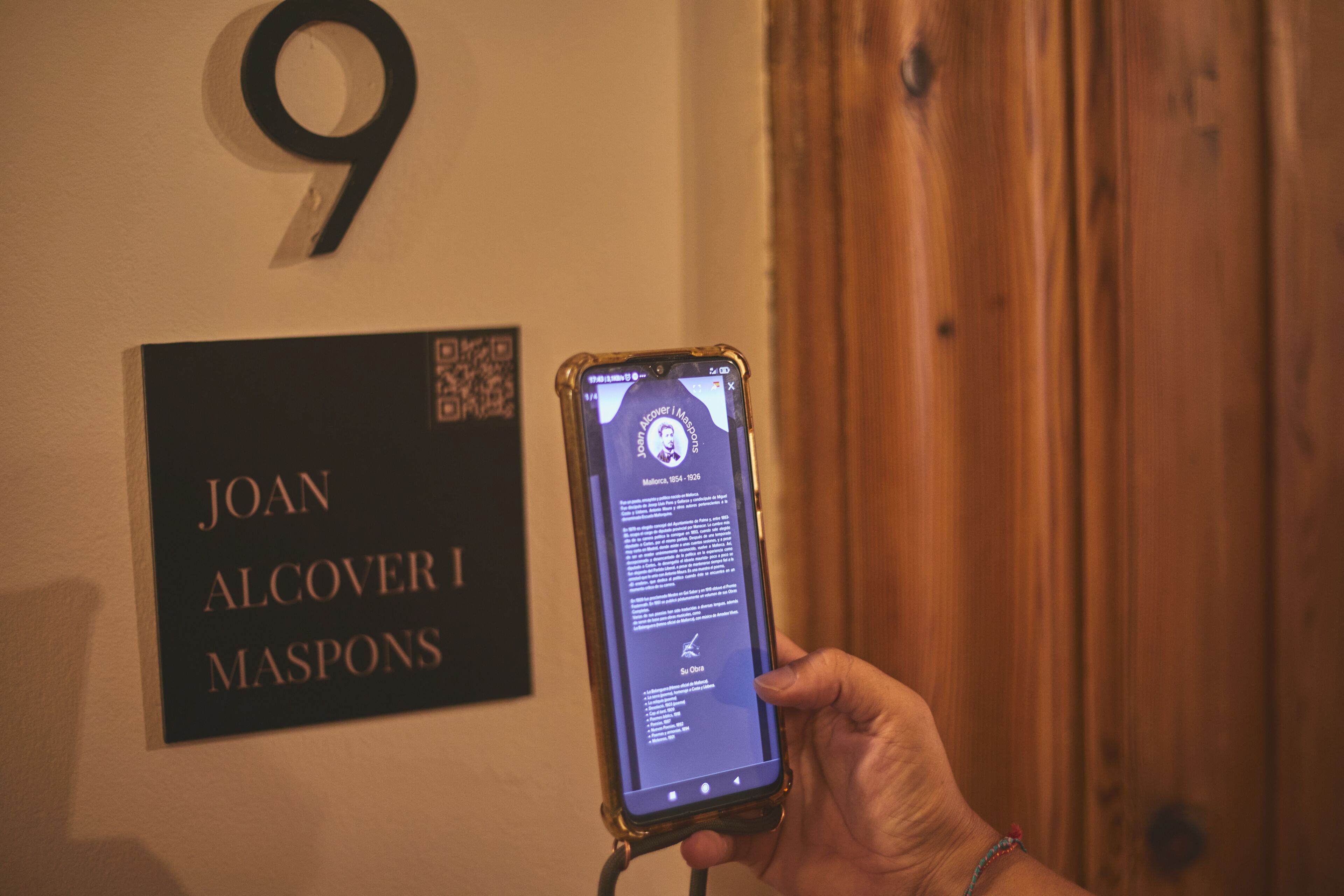 Una mano sosteniendo un teléfono inteligente que muestra un código QR. En la pared, se ve un letrero con el nombre JOAN ALCOVER MASPONS.