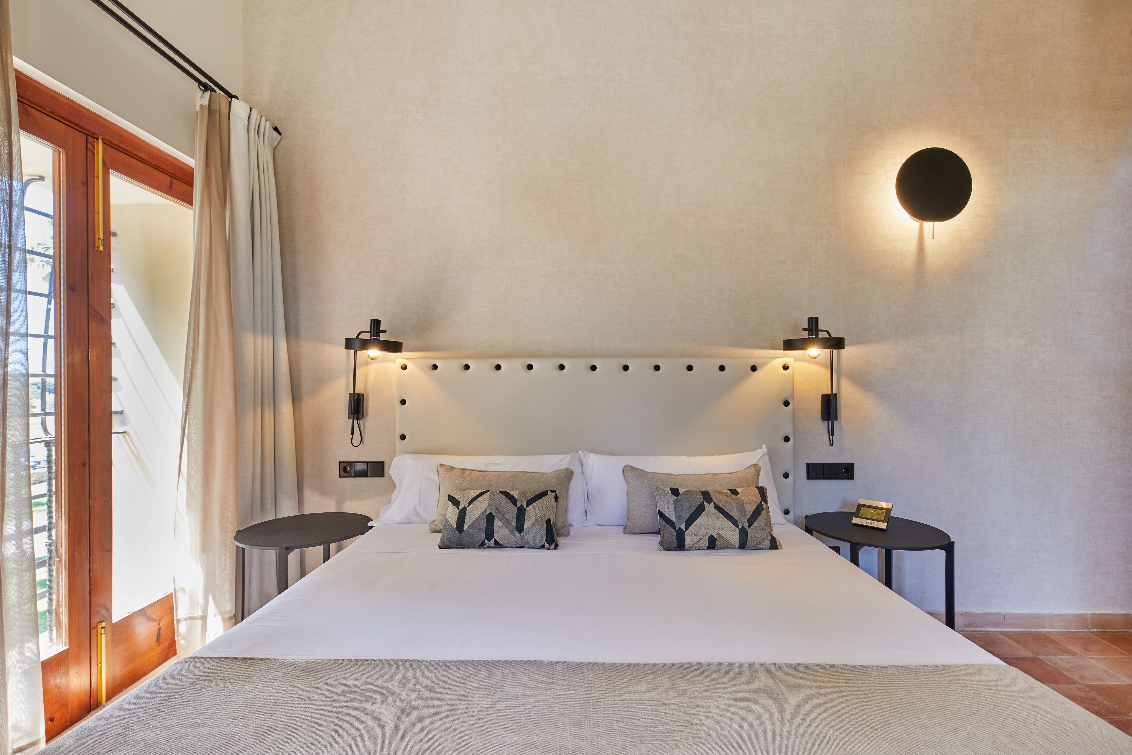 A bedroom with a large bed, two bedside tables, a wall-mounted light, and a view of the outdoors through a window with curtains.