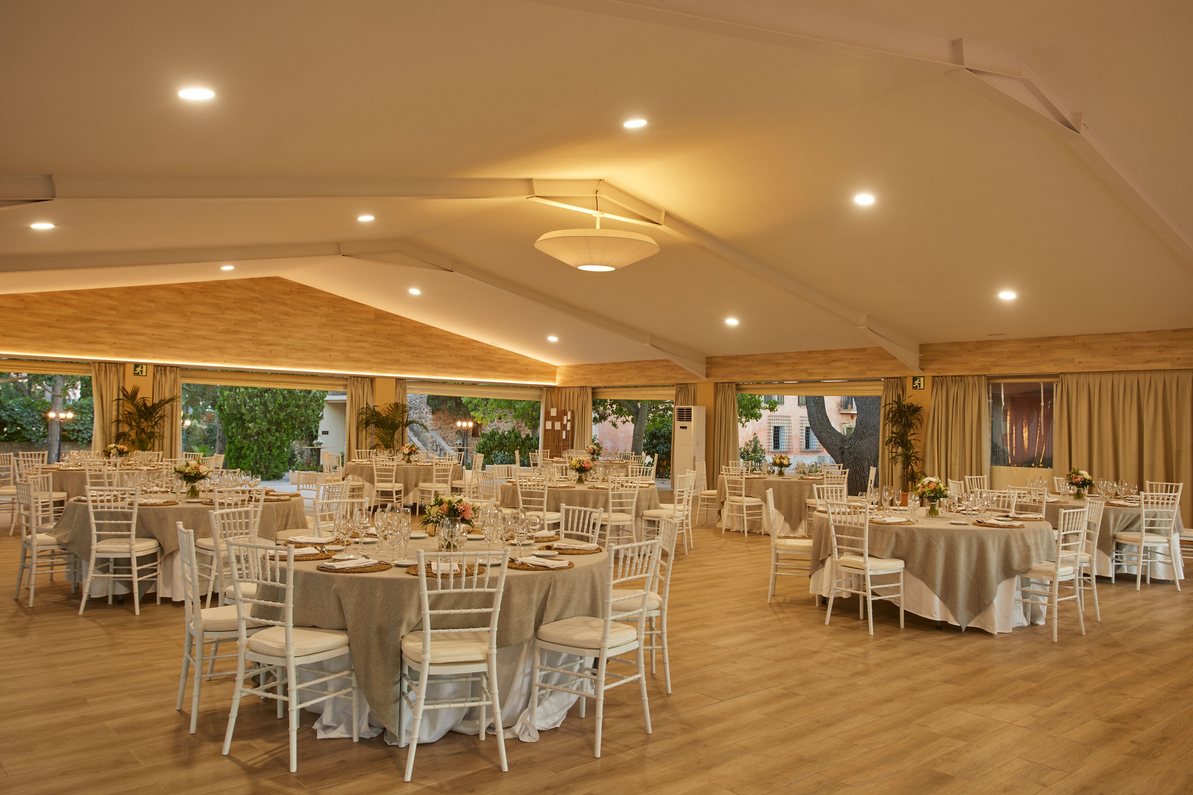 Une salle de banquet spacieuse, dotée de nombreuses tables rondes, de chaises blanches et de nappes beiges.