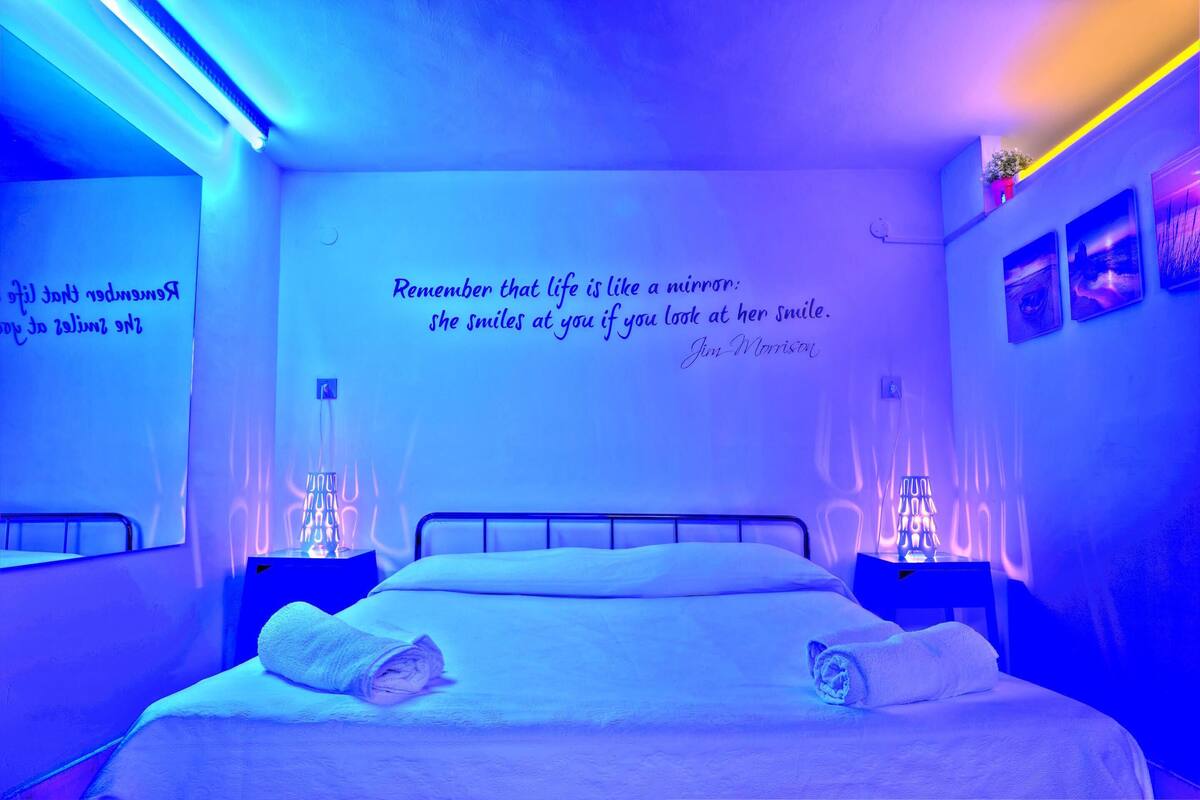 Un dormitorio con cama, dos mesitas de noche, una pared con una cita y un estante en la pared con una planta.