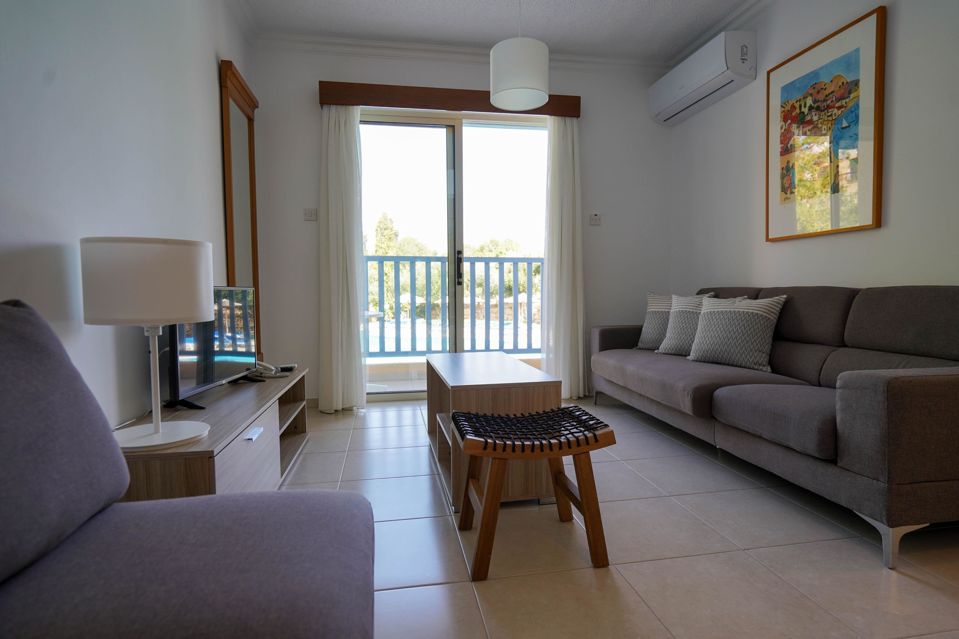 A living room with a sofa, a chair, a TV, a lamp, and a balcony with a view.