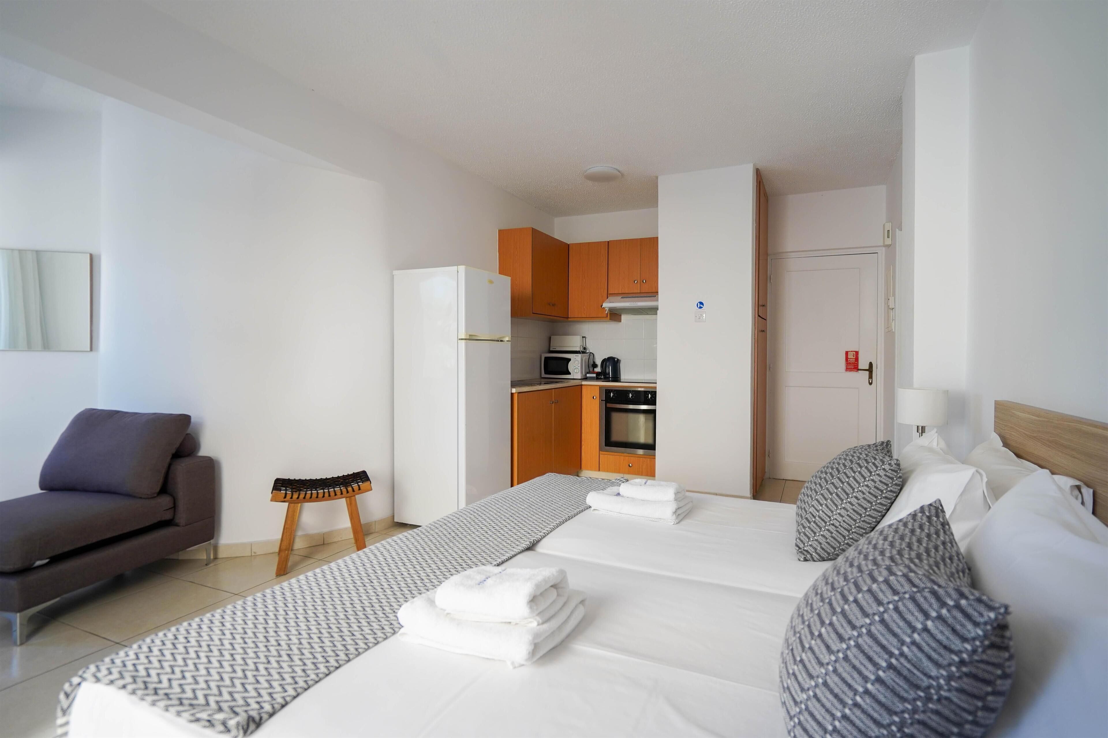 A hotel room with two beds, a sofa, a small stool, a kitchenette, and a door leading to another room.