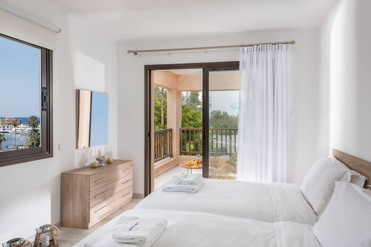 A bedroom with two beds, a wooden dresser, a balcony with a table and chairs, and a view of the sea.