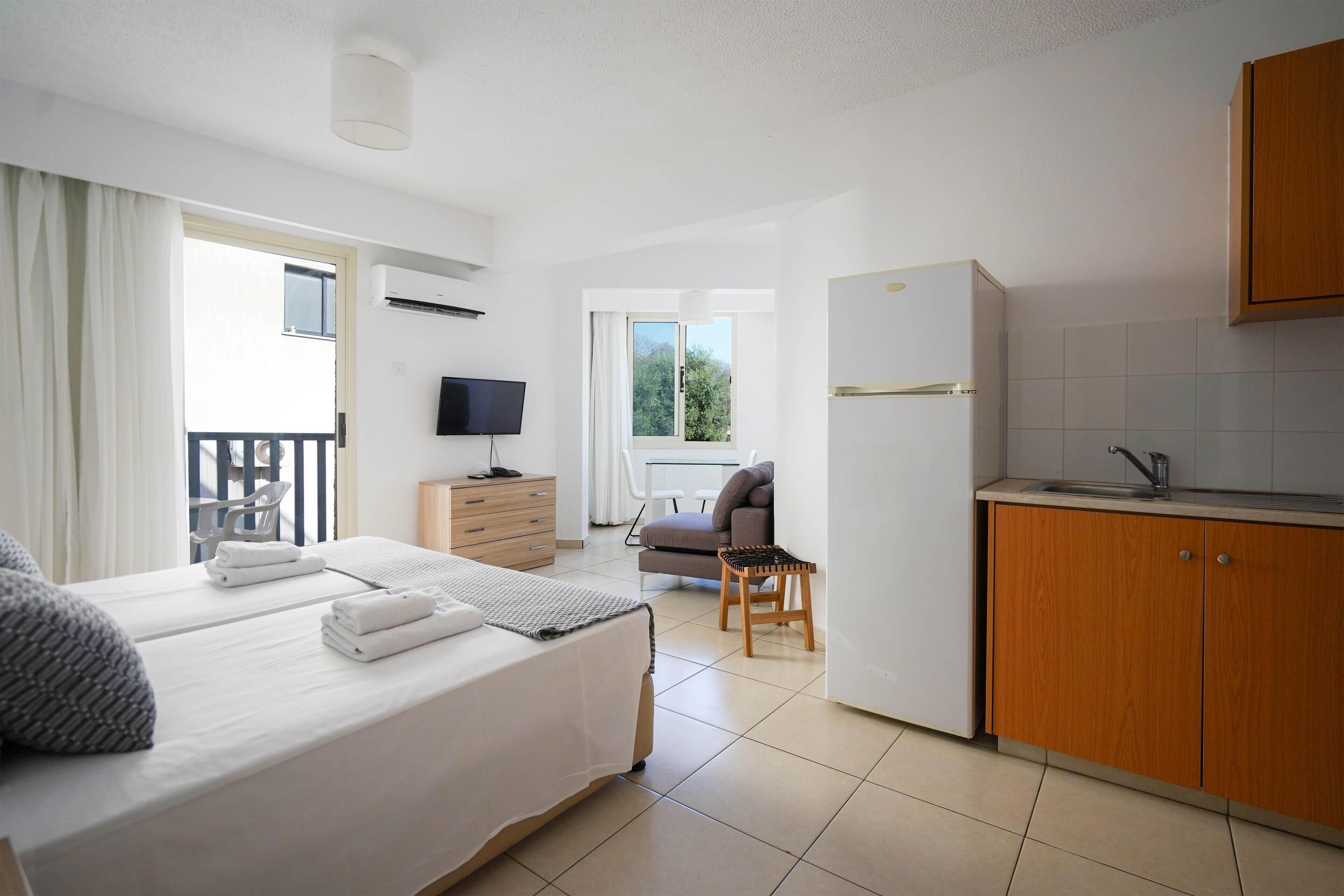 Quarto de hotel moderno com cama, kitchenette, televisão e varanda com cadeira e mesa.