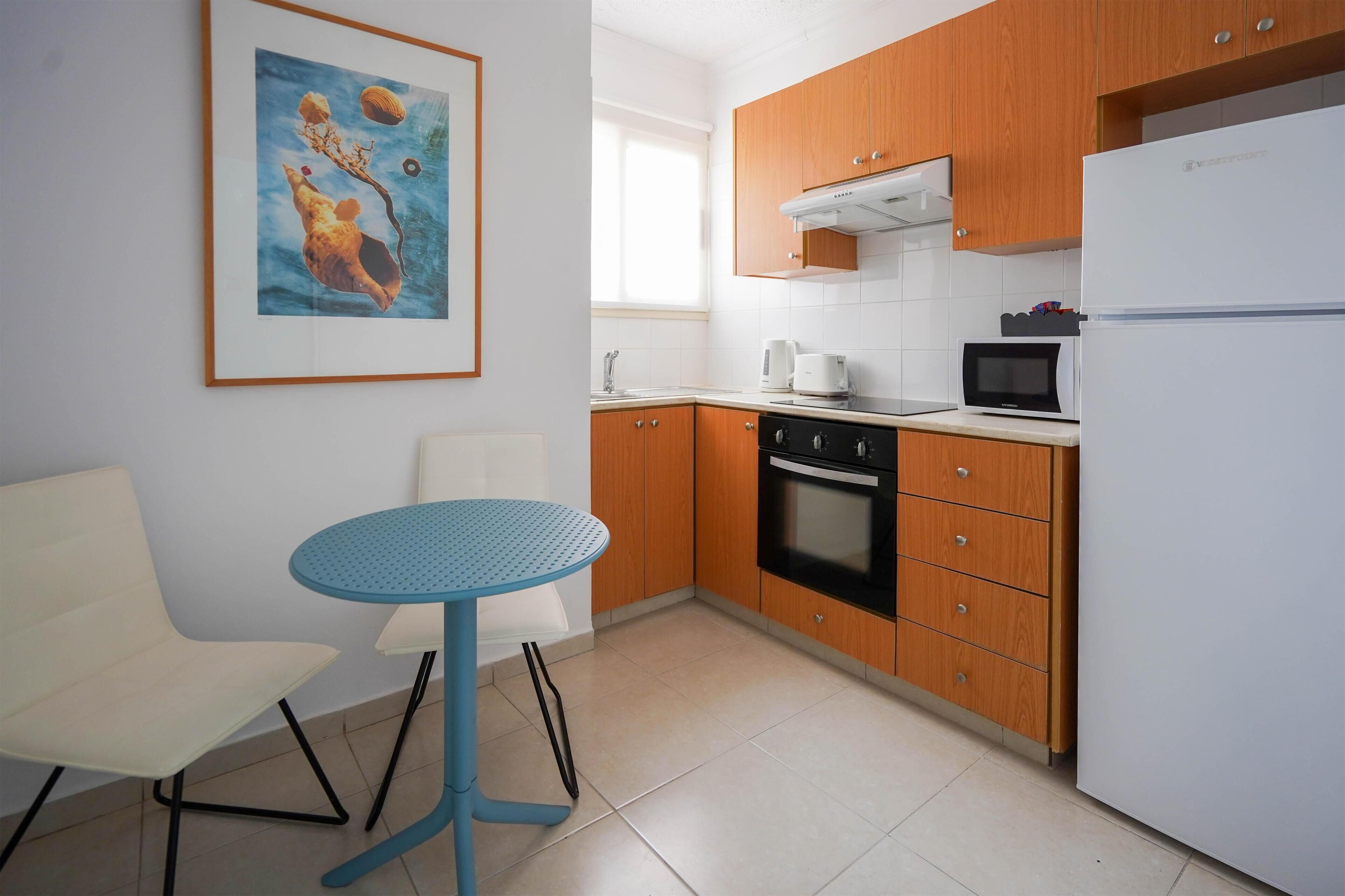 A compact kitchen with wooden cabinets, a white refrigerator, a microwave, and a coffee maker. There is a blue table and a white chair.