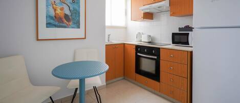 A compact kitchen with wooden cabinets, a white refrigerator, a microwave, and a coffee maker. There is a blue table and a white chair.