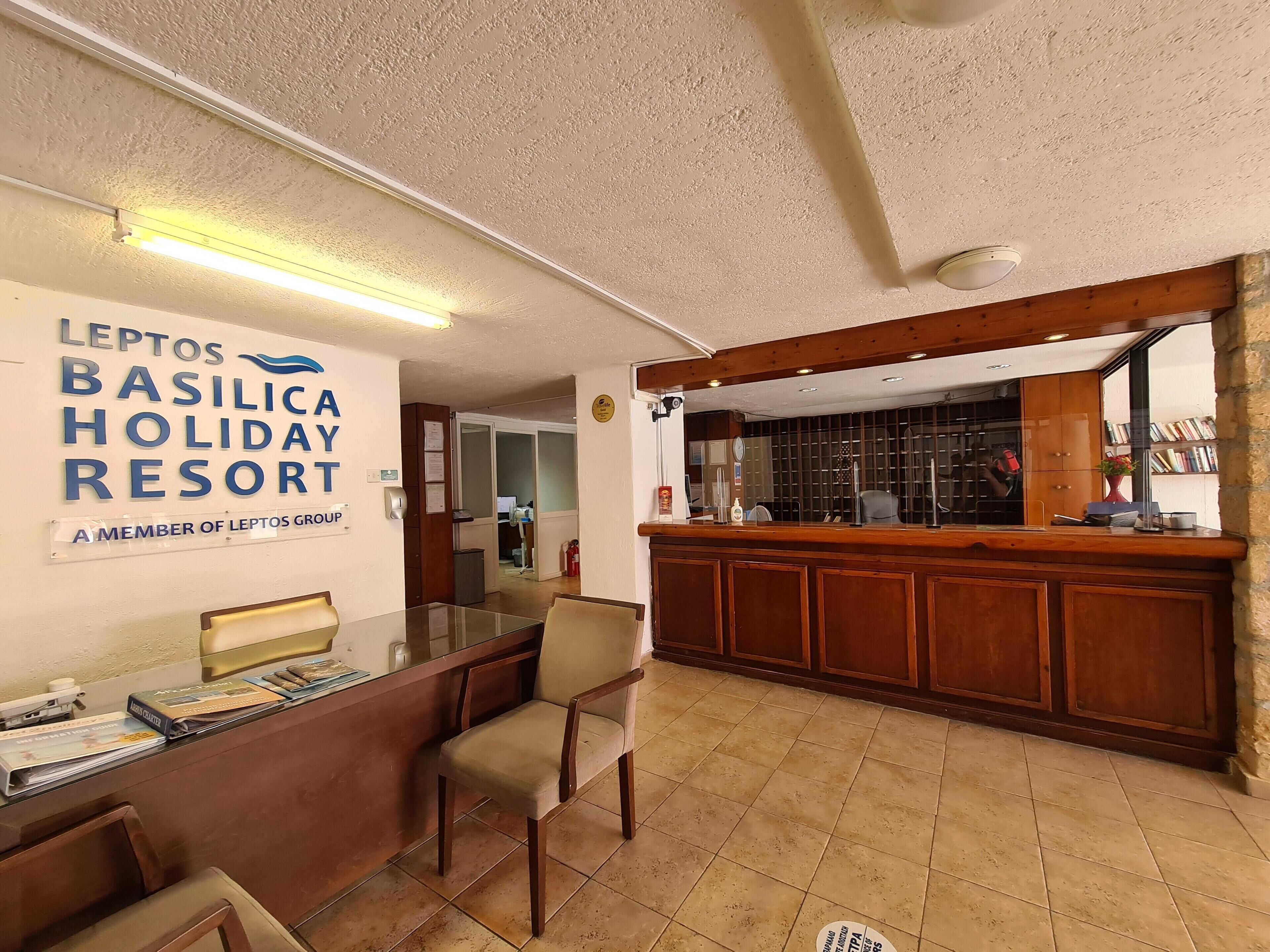Zona de recepción con un mostrador de madera, un escritorio de cristal y un cartel en la pared para Leptos Basilica Holiday Resort.