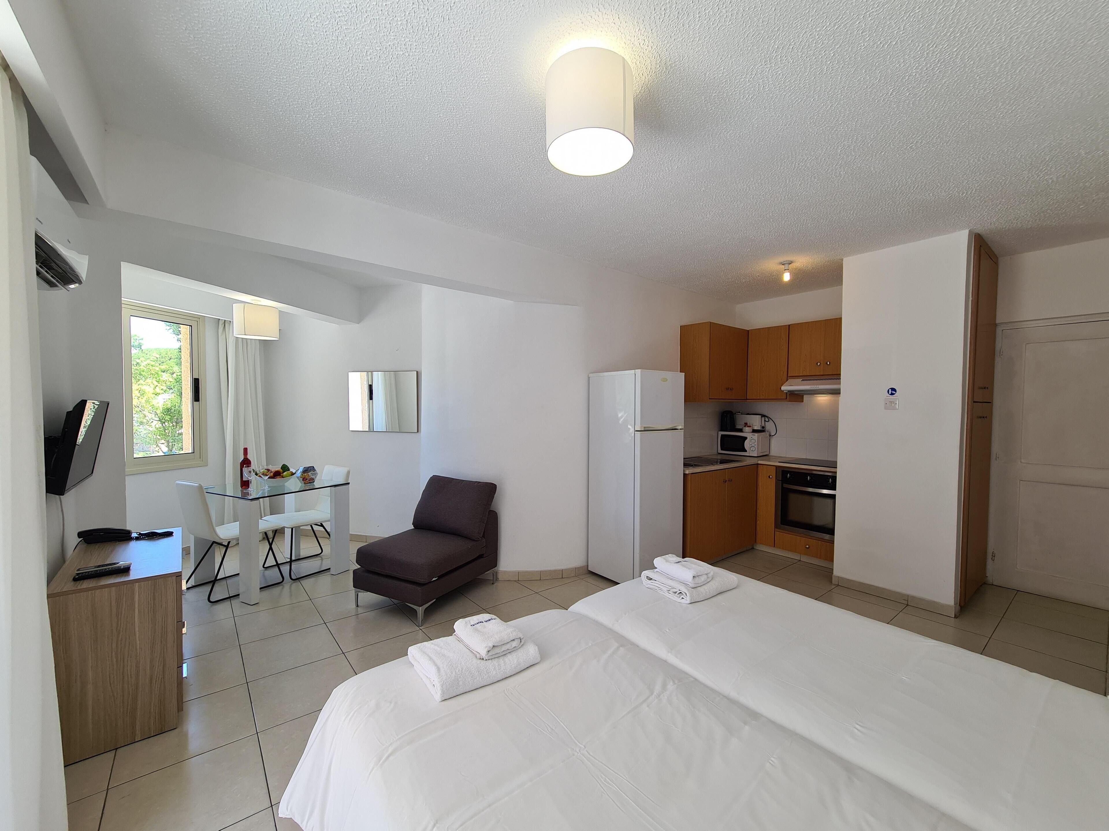A hotel room with a bed, a desk, a chair, a TV, a kitchen area, and a bathroom.