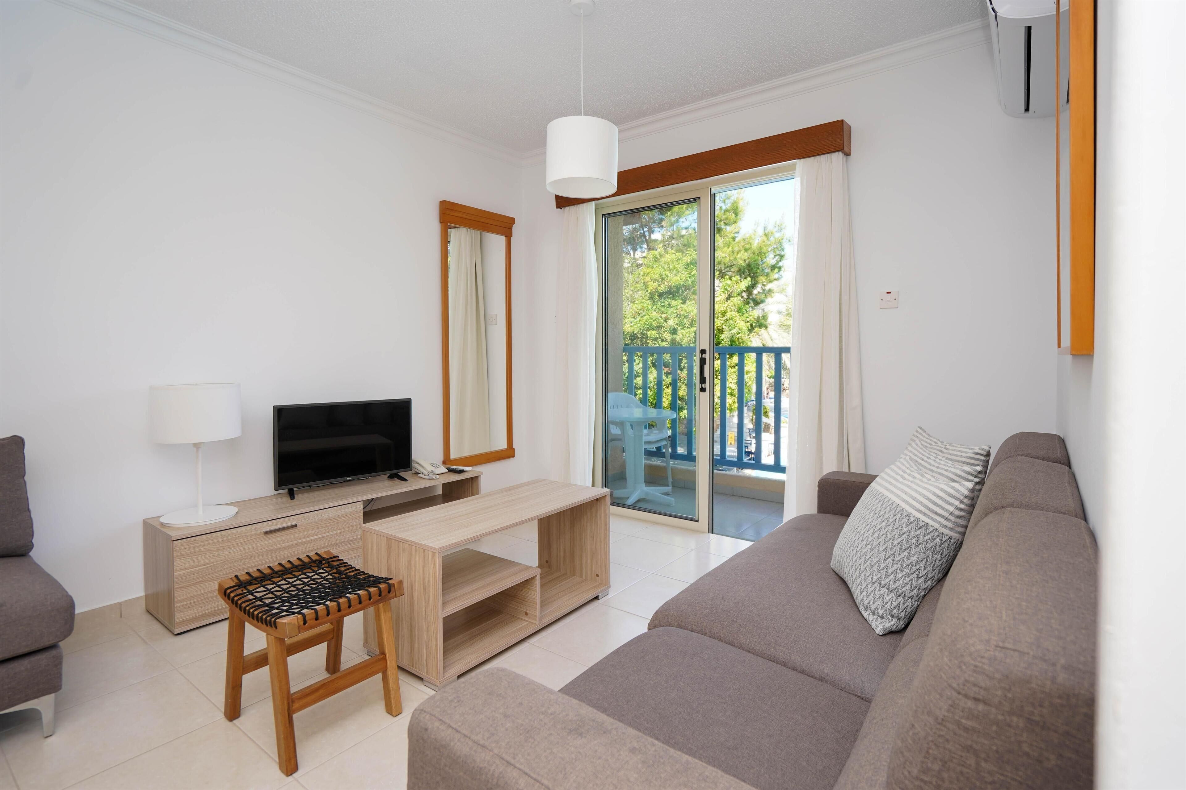 Un salon moderne avec un canapé, un téléviseur, une table basse en bois et un balcon doté d’une porte vitrée.