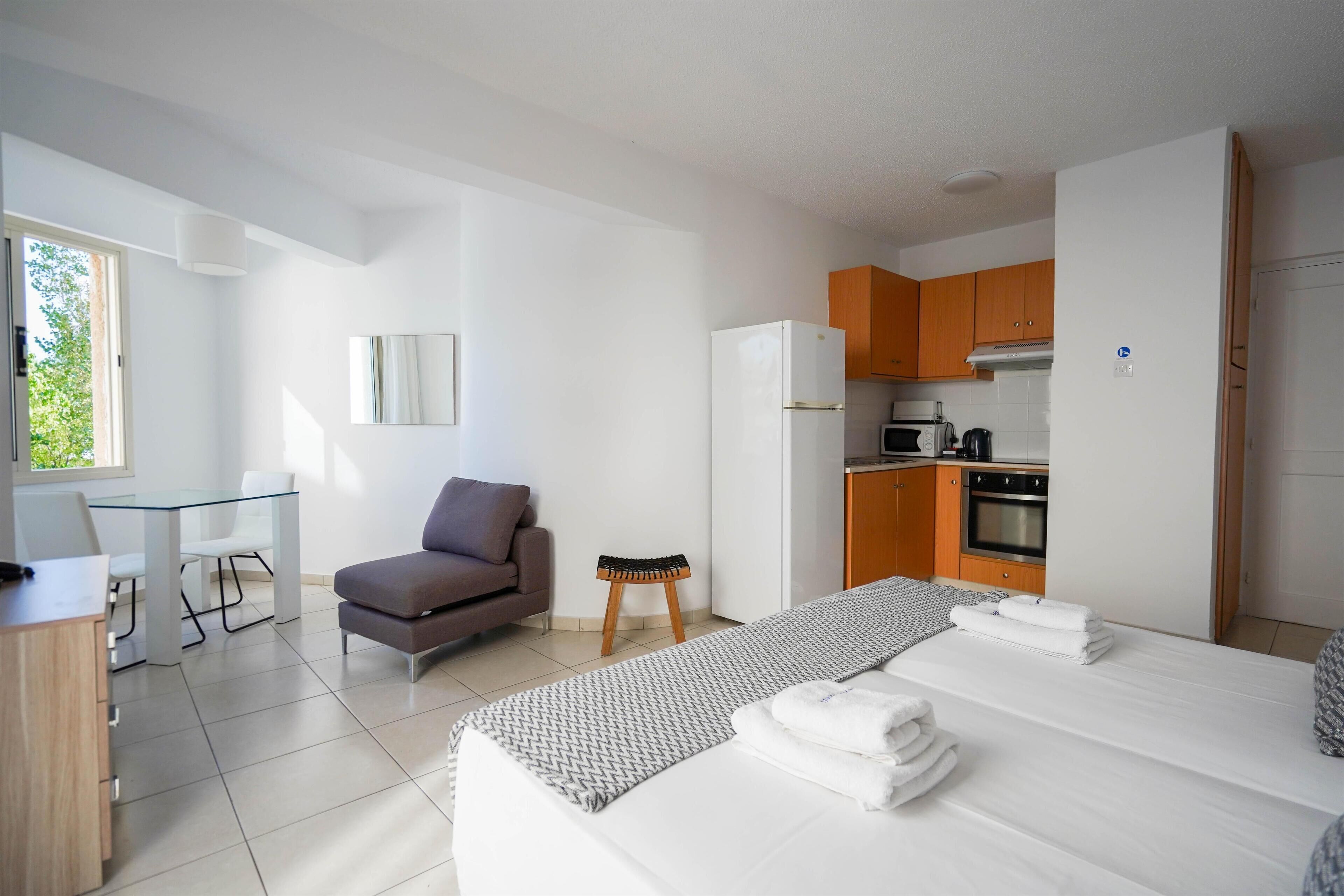 Una habitación de hotel moderna con cama, escritorio, silla, sofá, taburete pequeño y kitchenette.