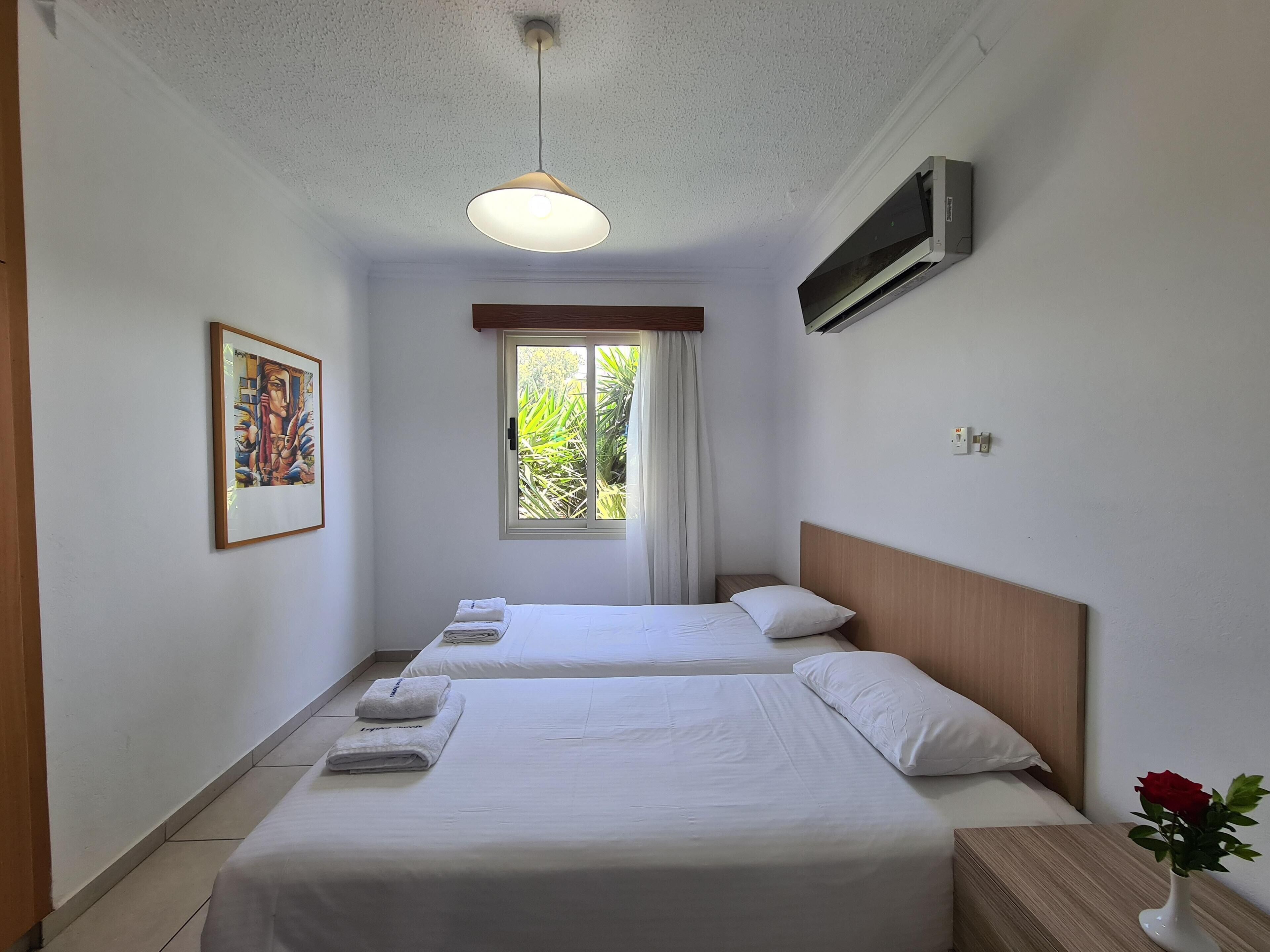 A hotel room with two beds, a window with curtains, a painting on the wall, and a vase with a rose on the nightstand.