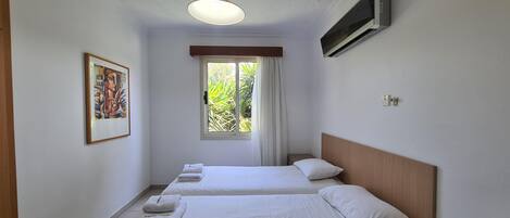 A hotel room with two beds, a window with curtains, a painting on the wall, and a vase with a rose on the nightstand.