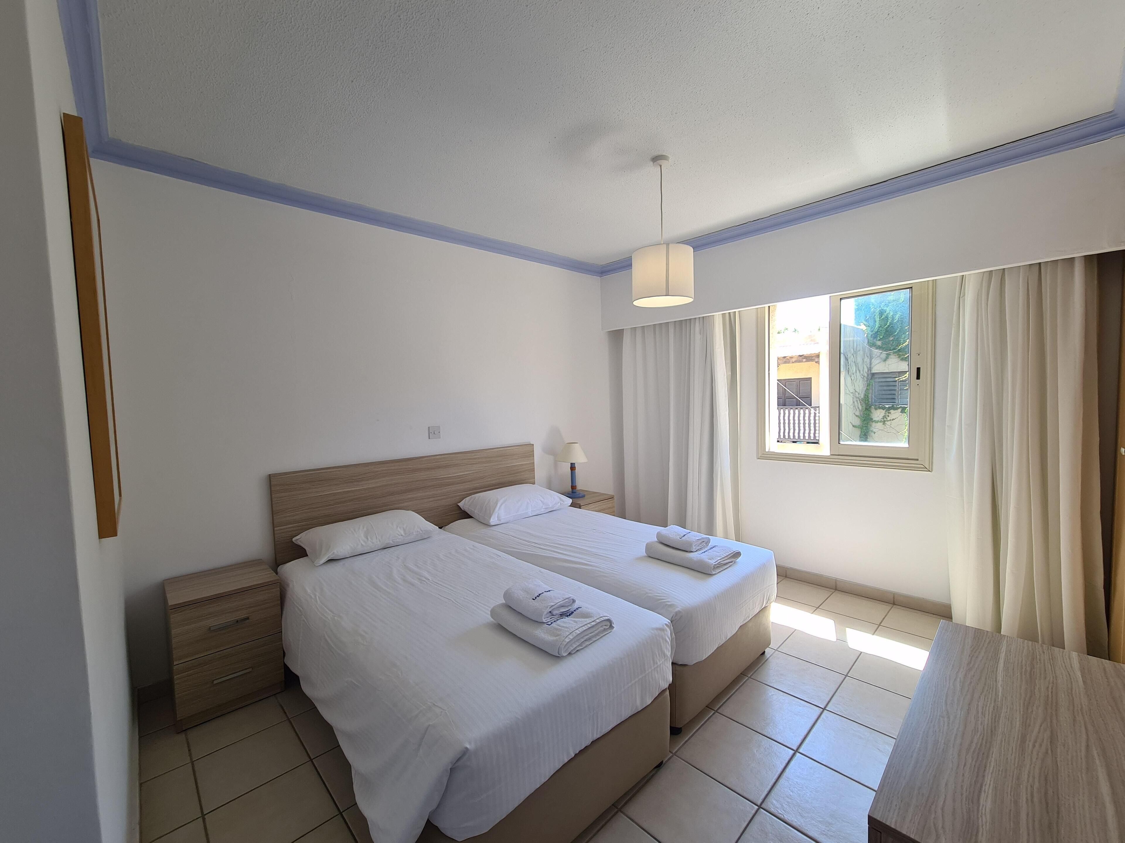 A hotel room with two beds, a wooden headboard, a nightstand, a lamp, and a window with curtains.