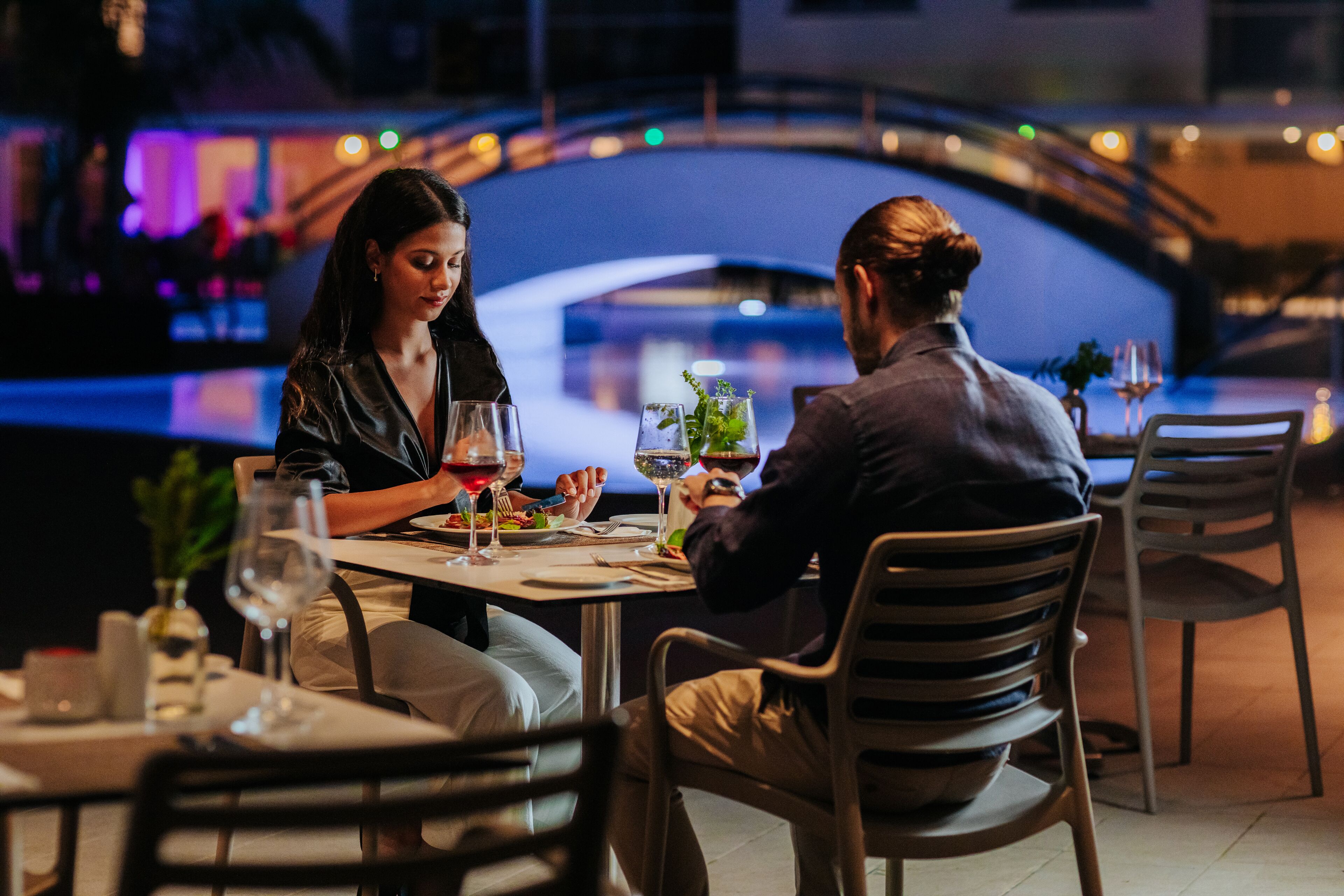 Una pareja cenando en una mesa con vista a un puente y al agua de fondo.