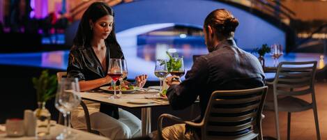 Una pareja cenando en una mesa con vista a un puente y al agua de fondo.