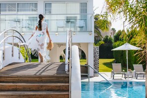 Zona de piscina con escalones que conducen a un balcón, sillas de descanso y vistas de un edificio moderno con barandillas de vidrio.