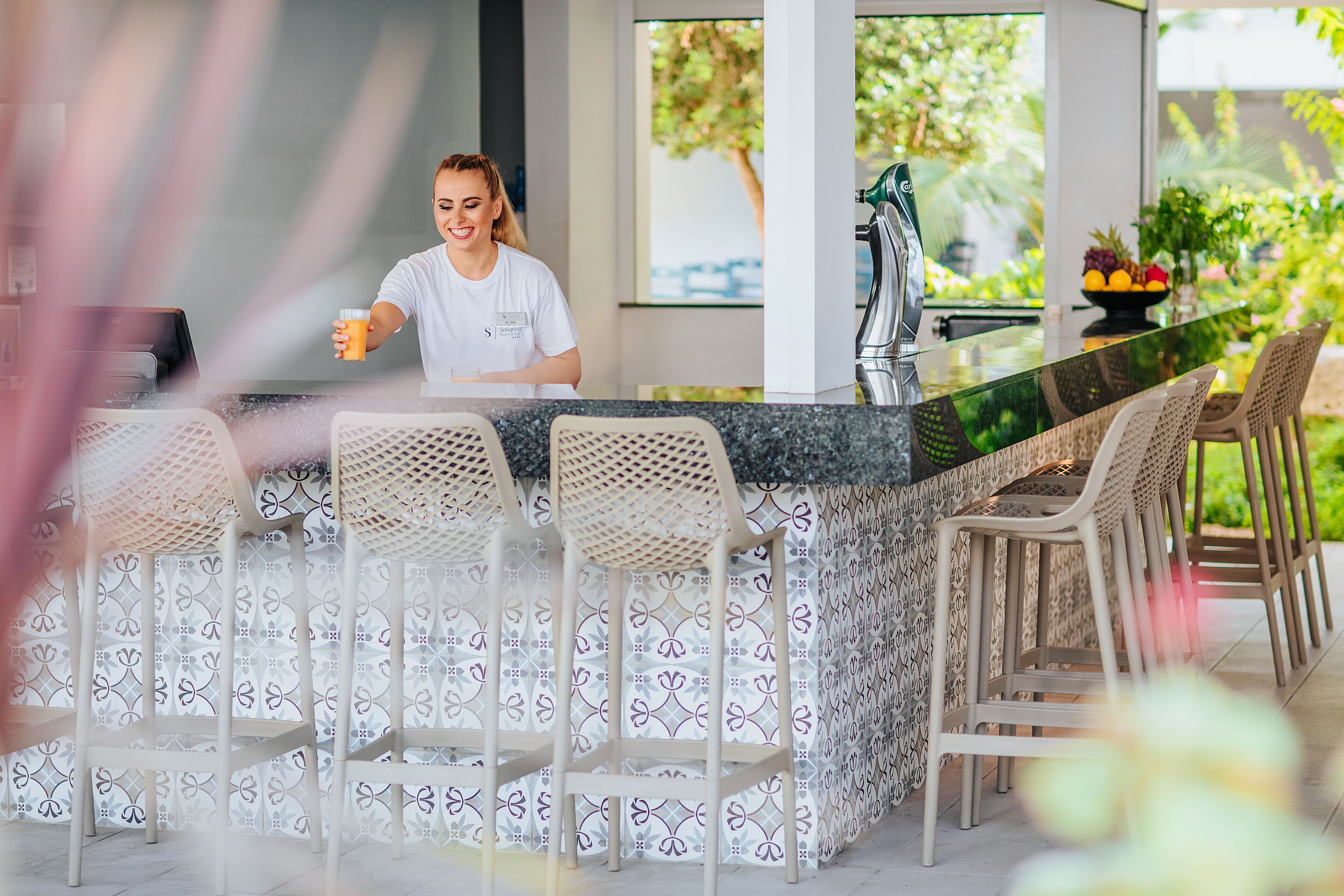 A modern kitchen with a bar area, white patterned chairs, and a view of greenery outside.