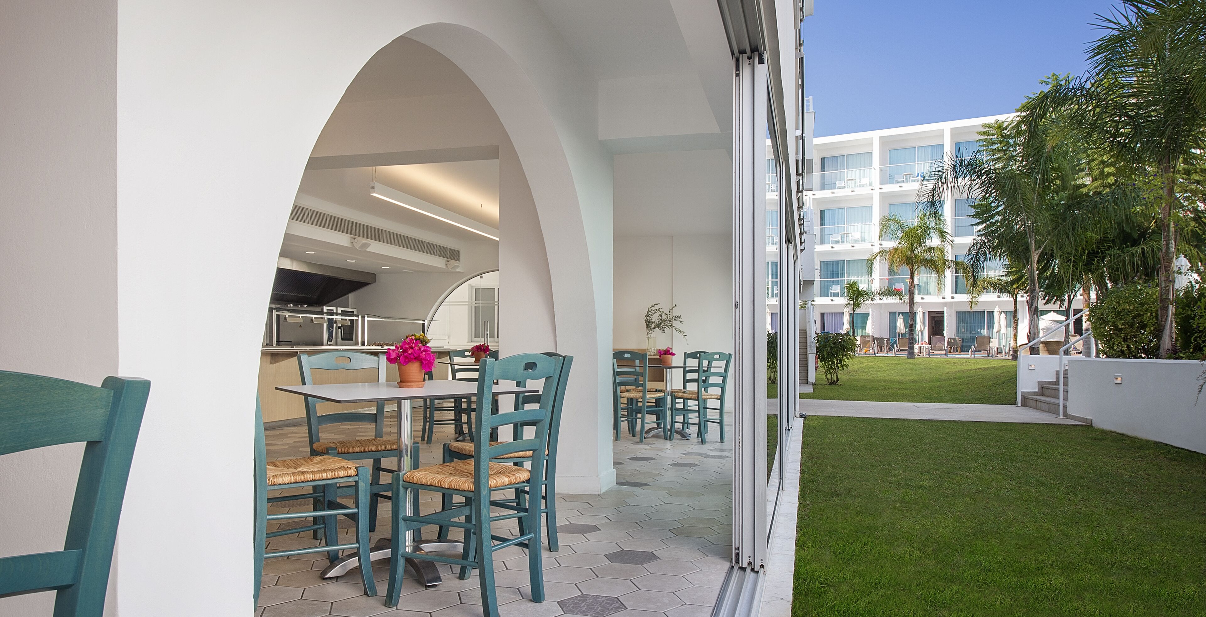 An outdoor dining area with a white table and teal chairs, a kitchen in the background, and a building with balconies and palm trees.