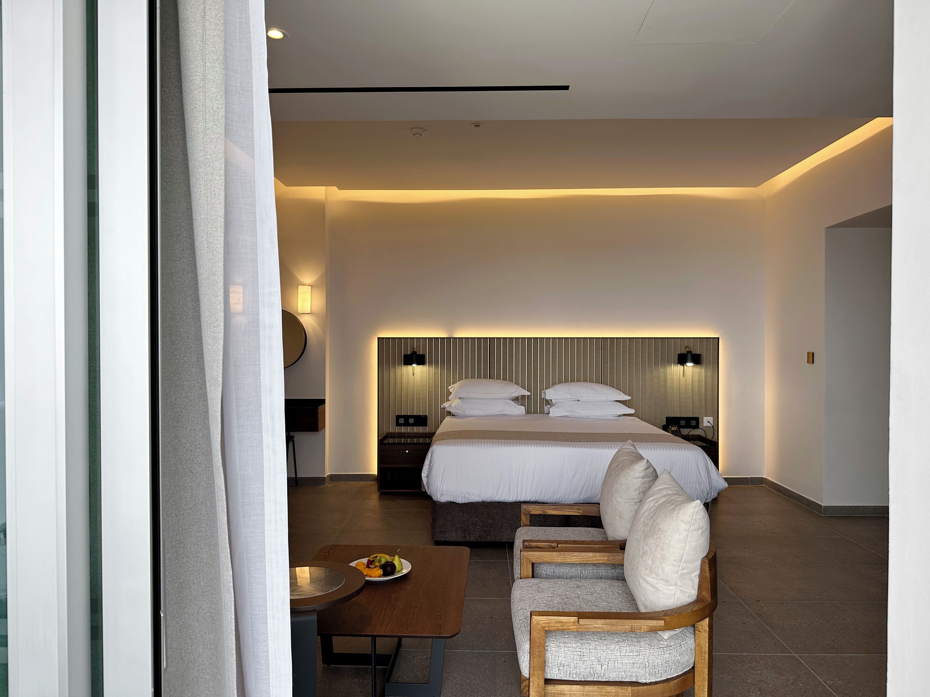 A modern hotel room with a large bed, bedside tables, a seating area with a table, and a wall-mounted lamp.