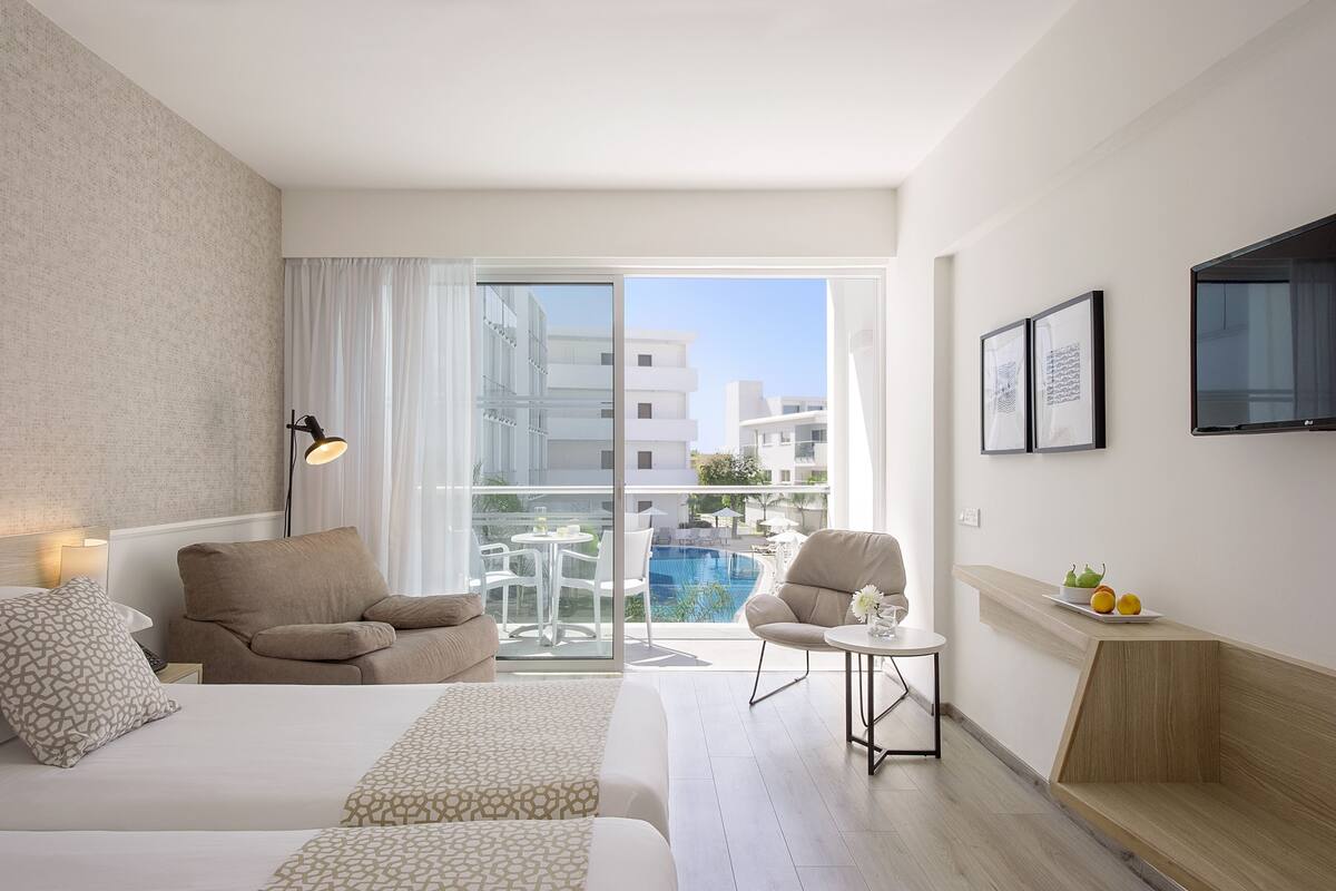 A hotel room with a bed, a sofa, a small table with a lamp, a TV on the wall, and a balcony with a pool and buildings in the background.