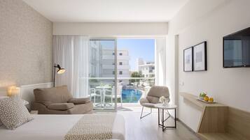 A hotel room with a bed, a sofa, a small table with a lamp, a TV on the wall, and a balcony with a pool and buildings in the background.