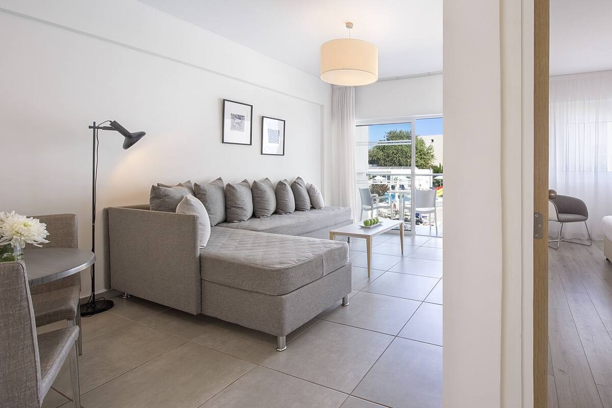 A modern living room with a grey sofa, a small table, and a floor lamp. There is a balcony with a view of the outside.