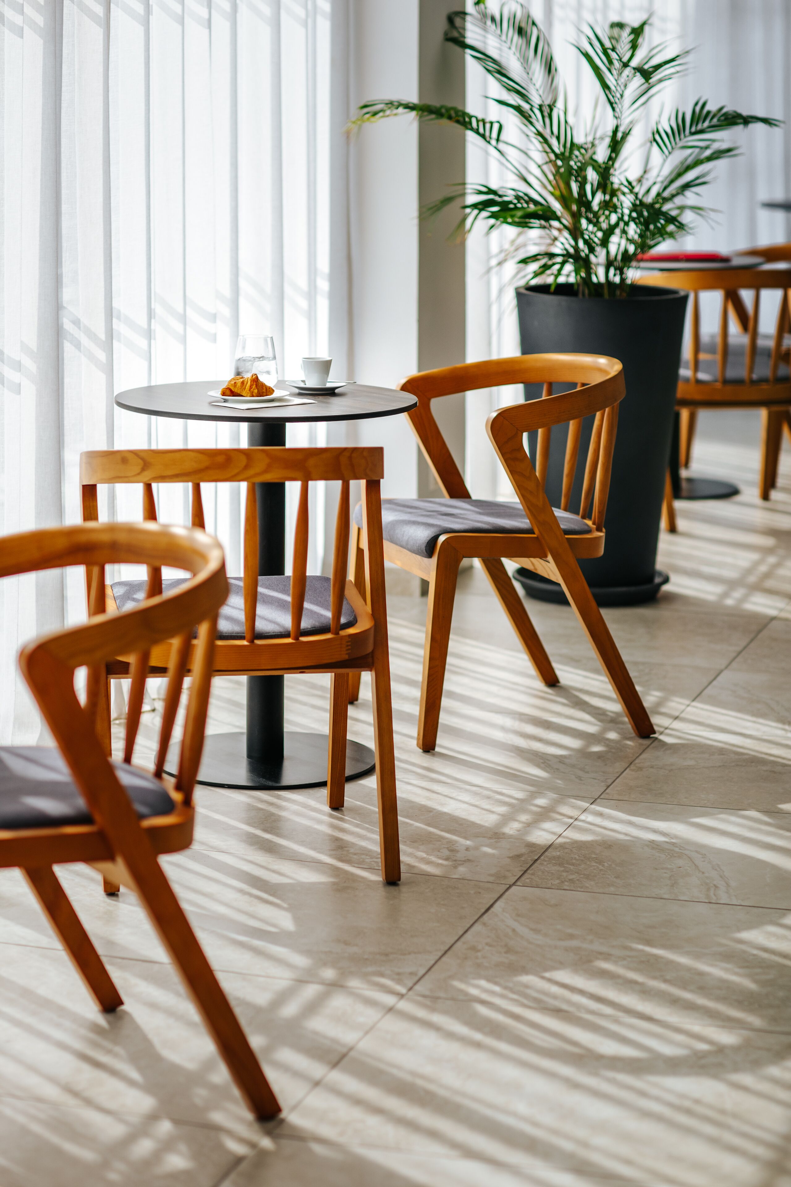 A seating area with wooden chairs and a round table, a potted plant, and a window with blinds.