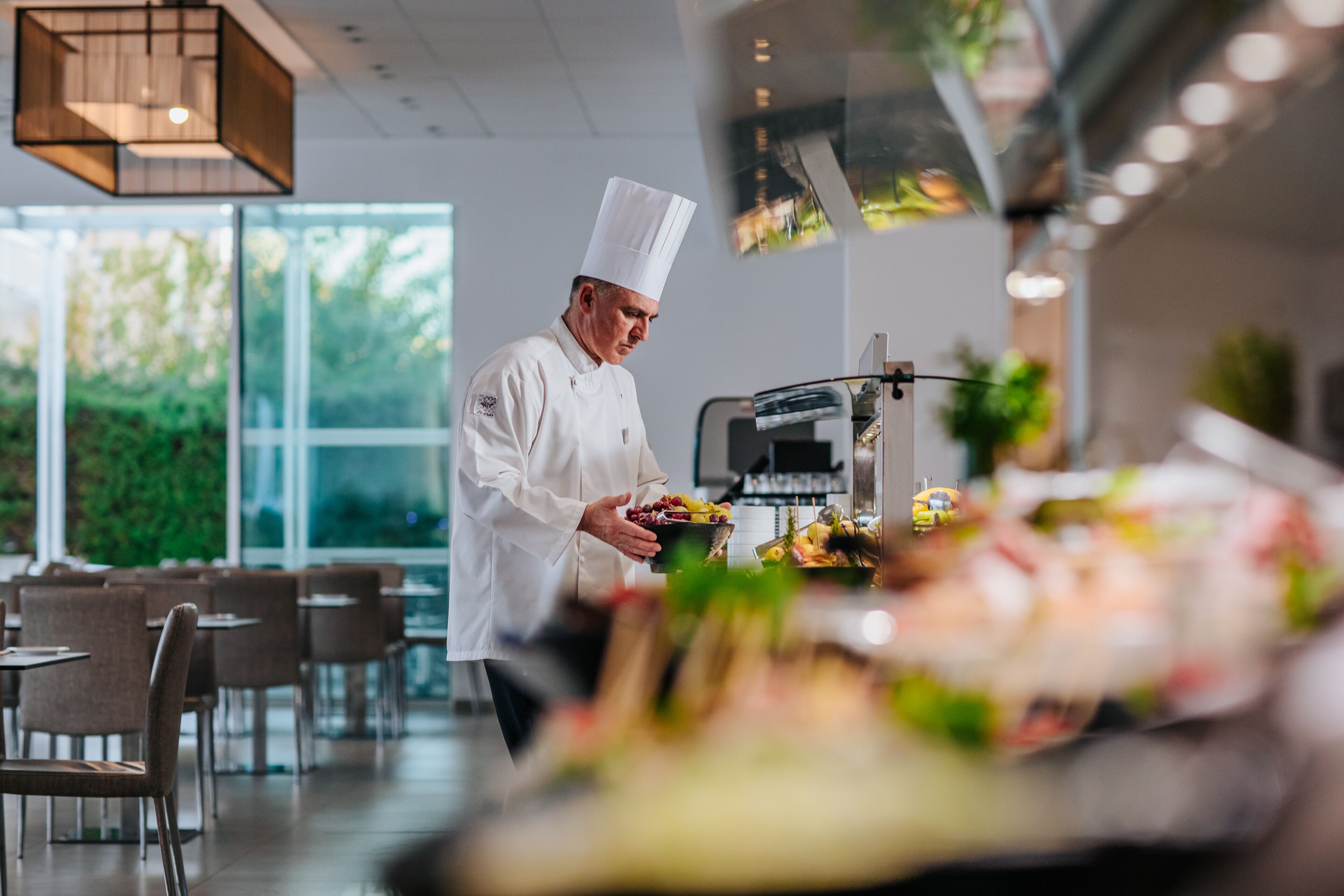 Un chef, vêtu d’un uniforme blanc et coiffé d’un haut chapeau, se tient dans un espace de restauration moderne, doté de grandes fenêtres, de tables et de chaises.