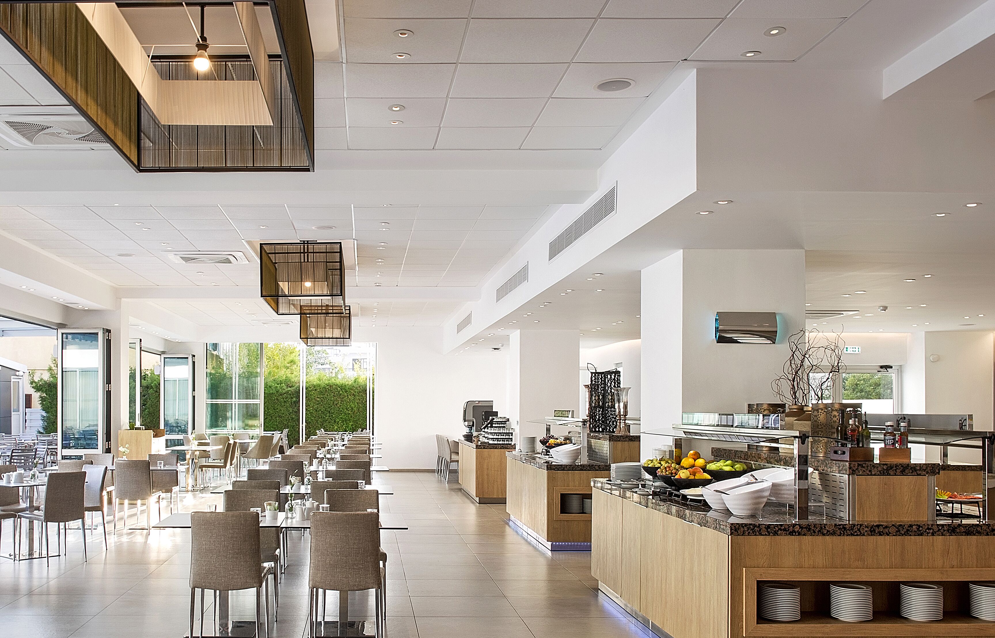 A modern dining area with multiple tables and chairs, a central counter with a fruit bowl, and a kitchen area visible in the background.