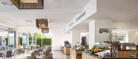 A modern dining area with multiple tables and chairs, a central counter with a fruit bowl, and a kitchen area visible in the background.