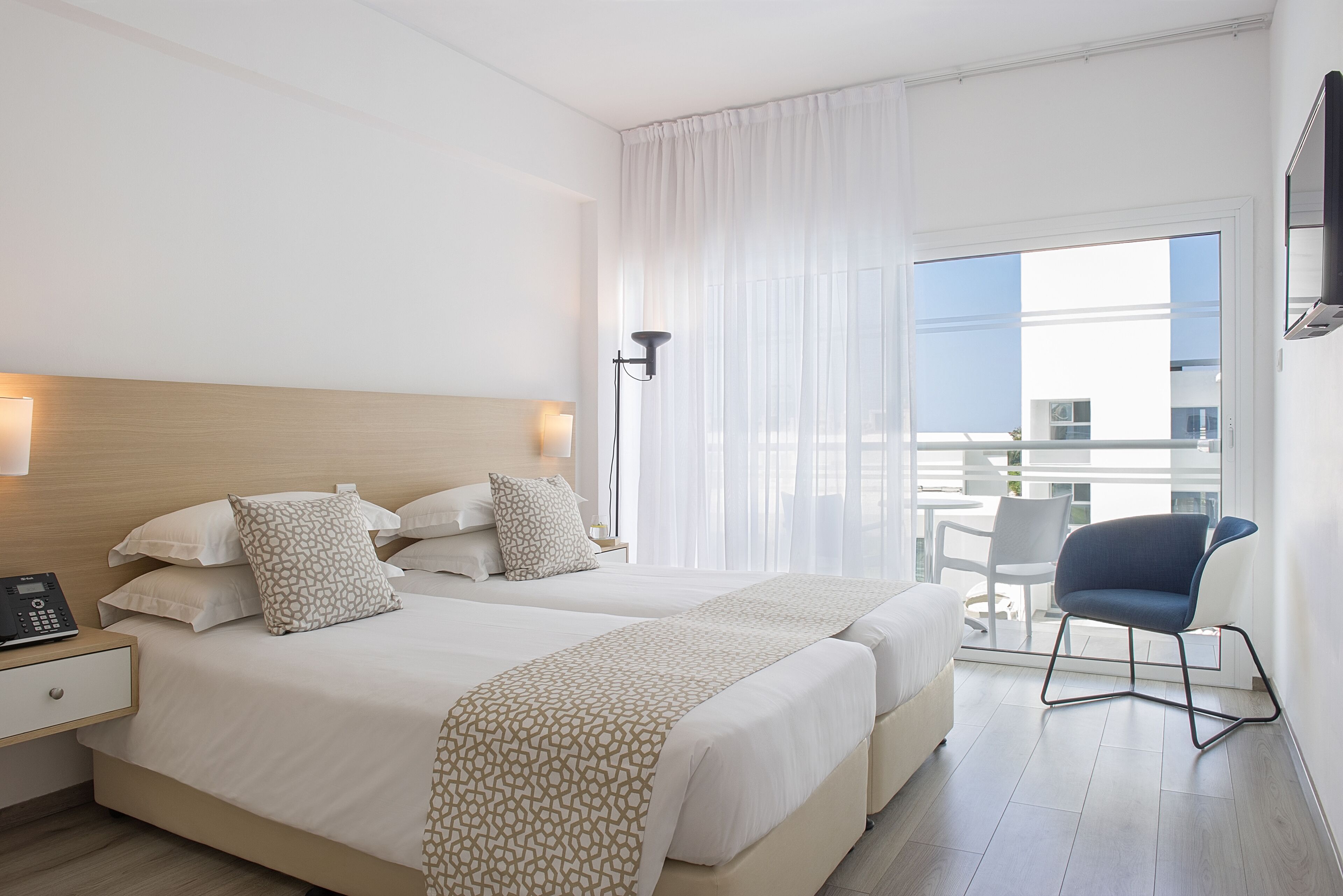 A hotel room with two beds, a bedside table with a phone, a chair, and a balcony with a table and chairs.