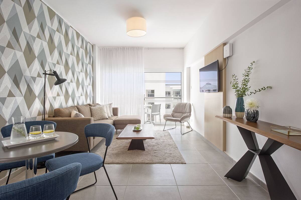 A modern living room with a sofa, coffee table, and chairs. There is a TV mounted on the wall and a plant on a side table.