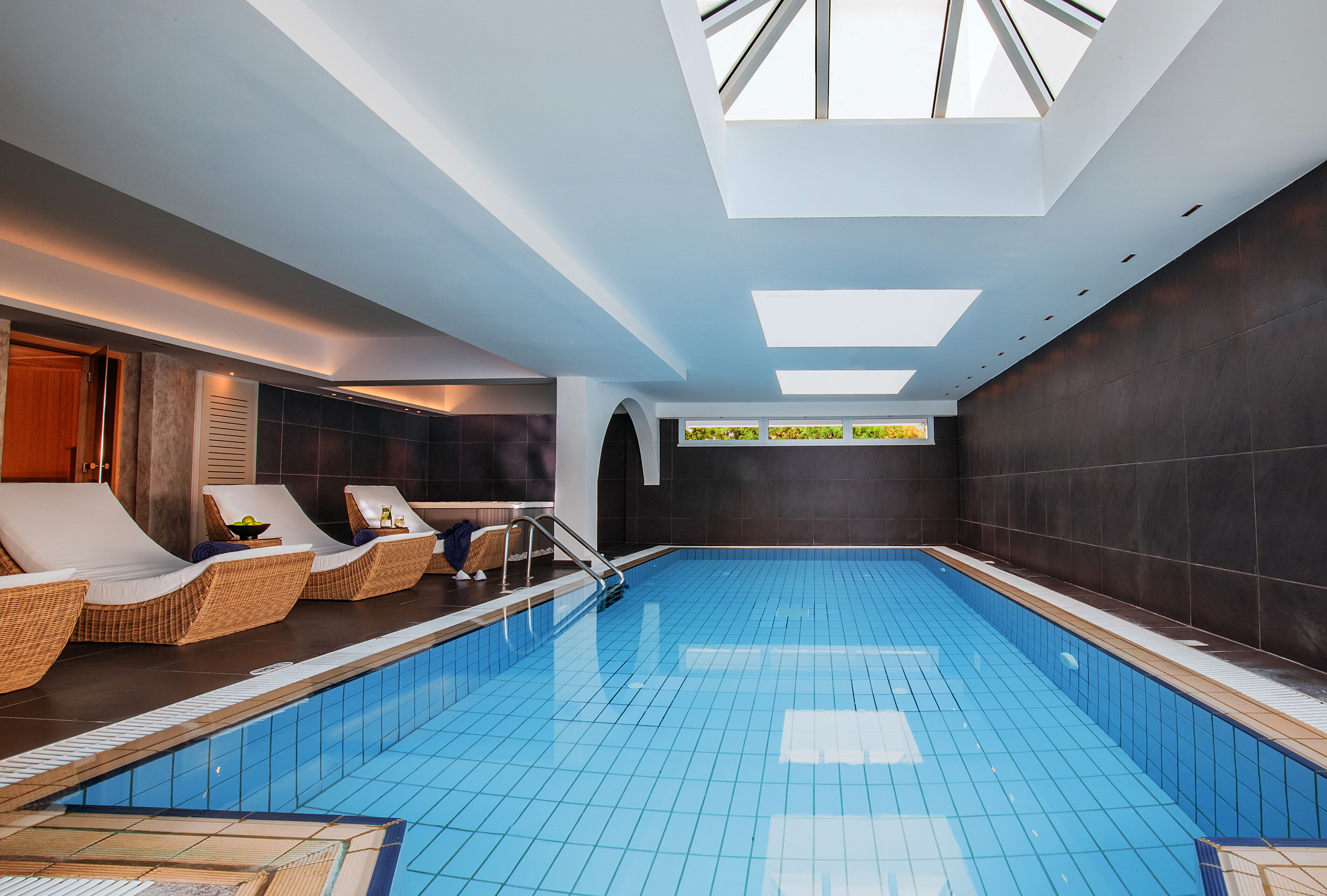 Une piscine intérieure moderne, dotée d’une verrière, de chaises longues et offrant une vue sur la verdure grâce à une fenêtre.