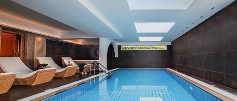 Une piscine intérieure moderne, dotée d’une verrière, de chaises longues et offrant une vue sur la verdure grâce à une fenêtre.