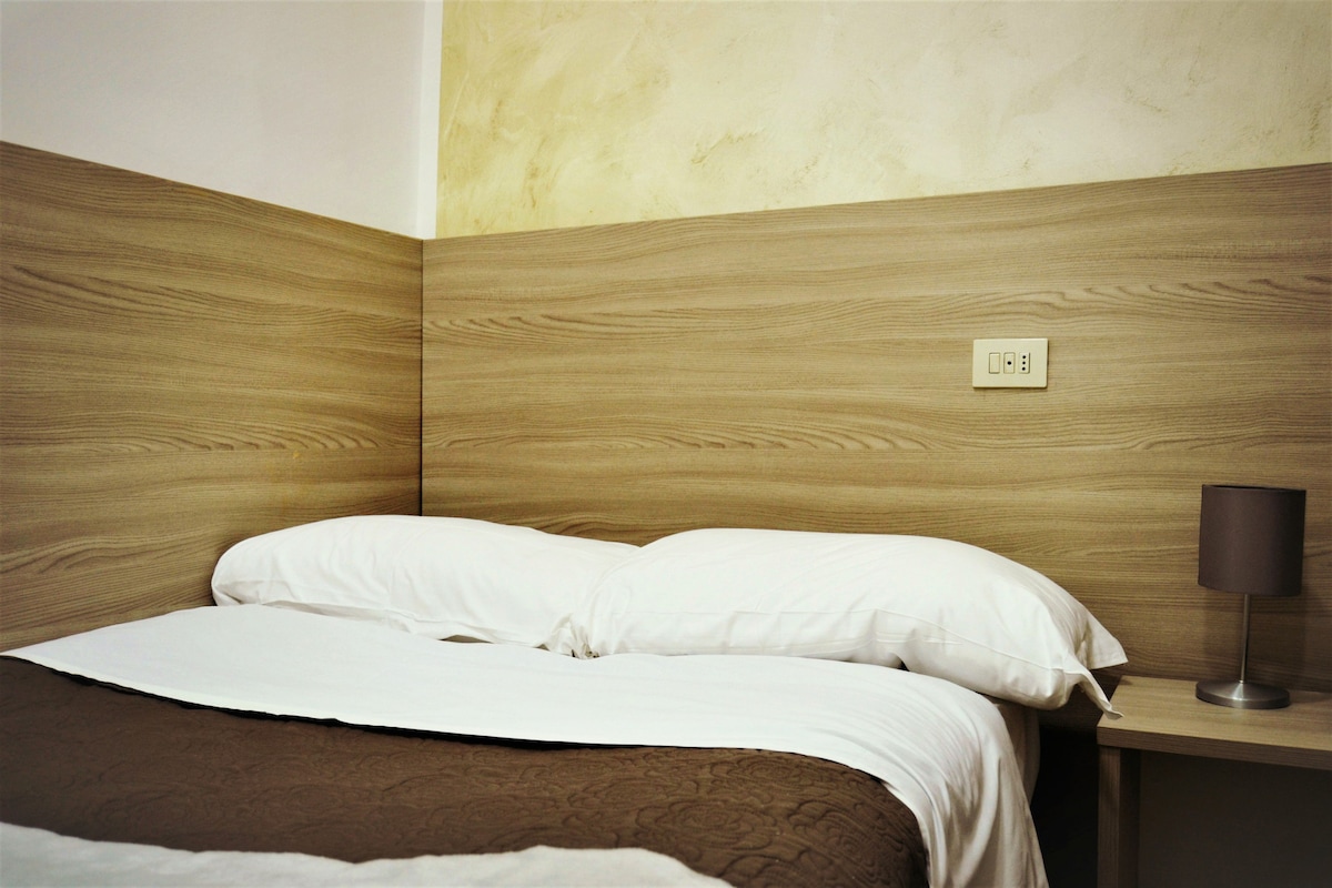 A neatly made bed with white linens and a brown throw blanket, a bedside lamp, and a power outlet on the wall.