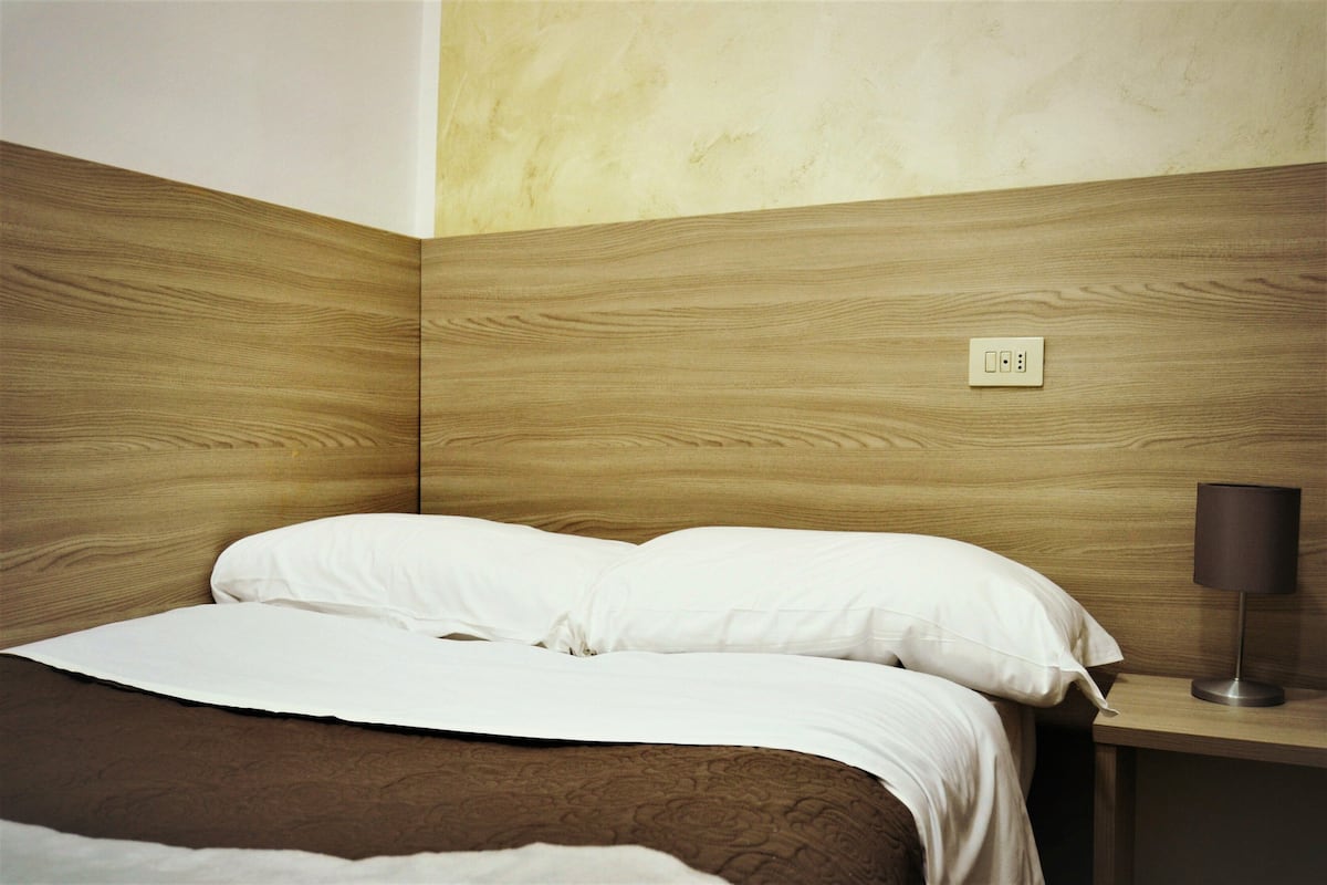 Un lit bien fait, avec du linge de lit blanc et une couverture marron, une lampe de chevet et une prise électrique sur le mur.