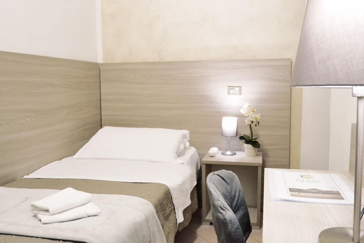Une chambre d’hôtel avec une tête de lit en bois, un lit recouvert de draps blancs, une table de chevet avec une lampe et un pot de fleurs, et une chaise.