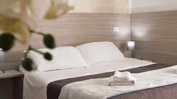 A hotel room with a wooden headboard, a white bedspread, and a neatly folded towel on the bed. - Hotel Colombo (Naples)