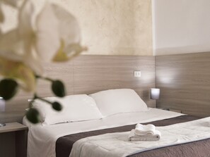 Habitación de hotel con cabecera de madera, colcha blanca y toalla doblada cuidadosamente sobre la cama.