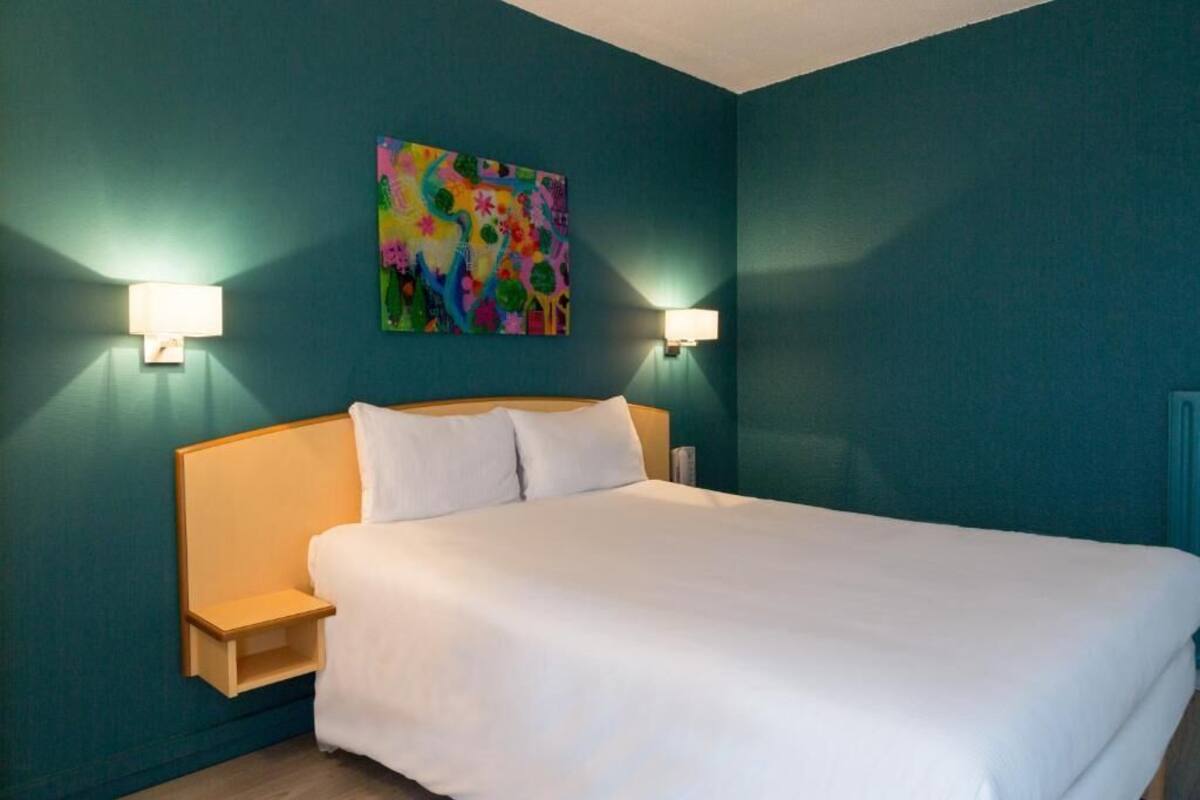 A hotel room with teal walls, a large bed with white bedding, two bedside lamps, and a colorful abstract painting above the bed.