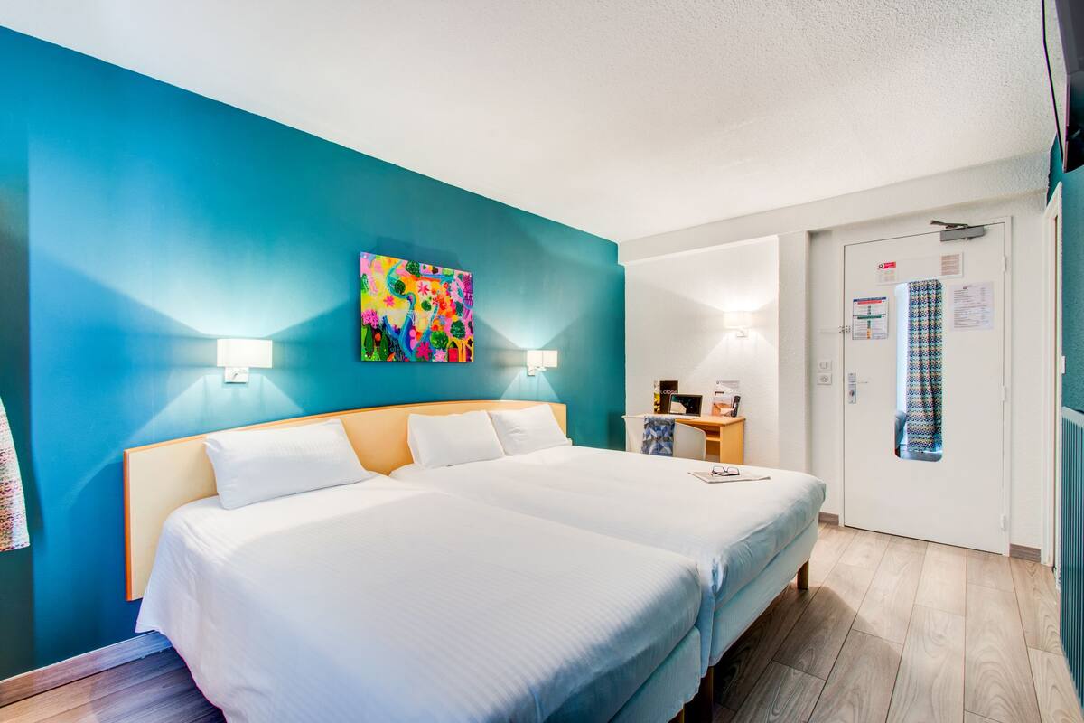 A hotel room with two beds, a colorful abstract painting on the wall, and a door with a patterned curtain.