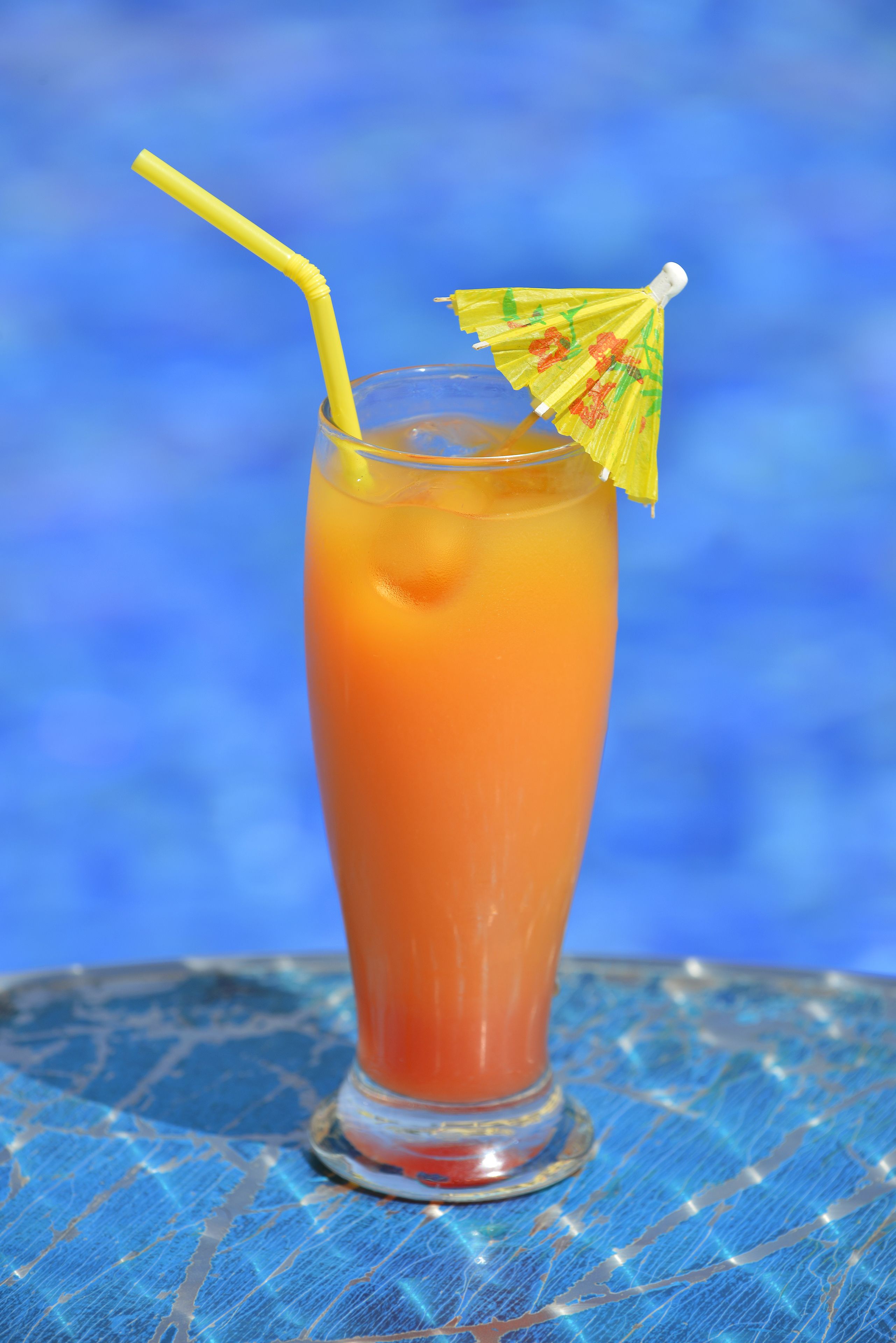 Ein hoher Glas mit einem gelben Strohhalm und einem kleinen gelben Regenschirm auf einer blauen, strukturierten Oberfläche.