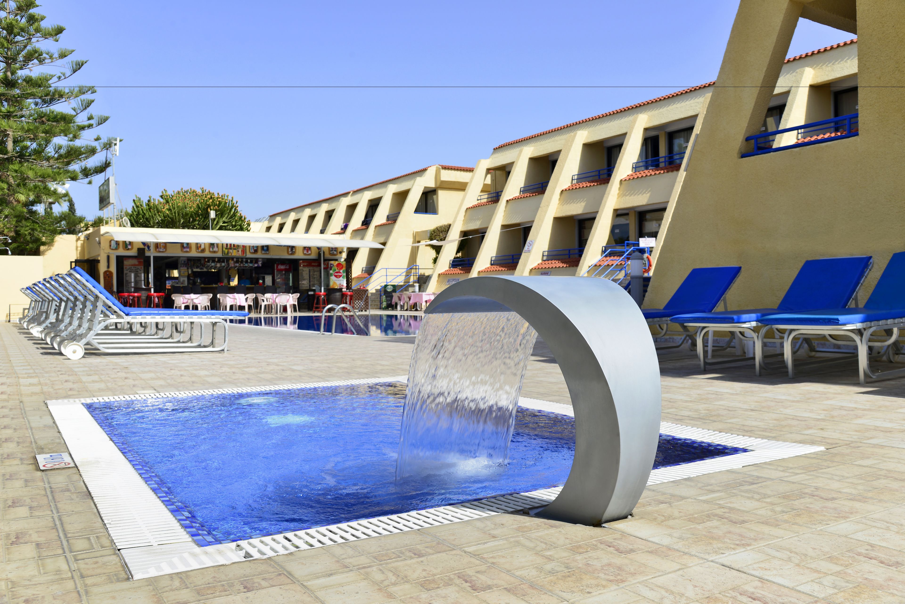 A swimming pool with a water feature, surrounded by lounge chairs and a building with multiple balconies.
