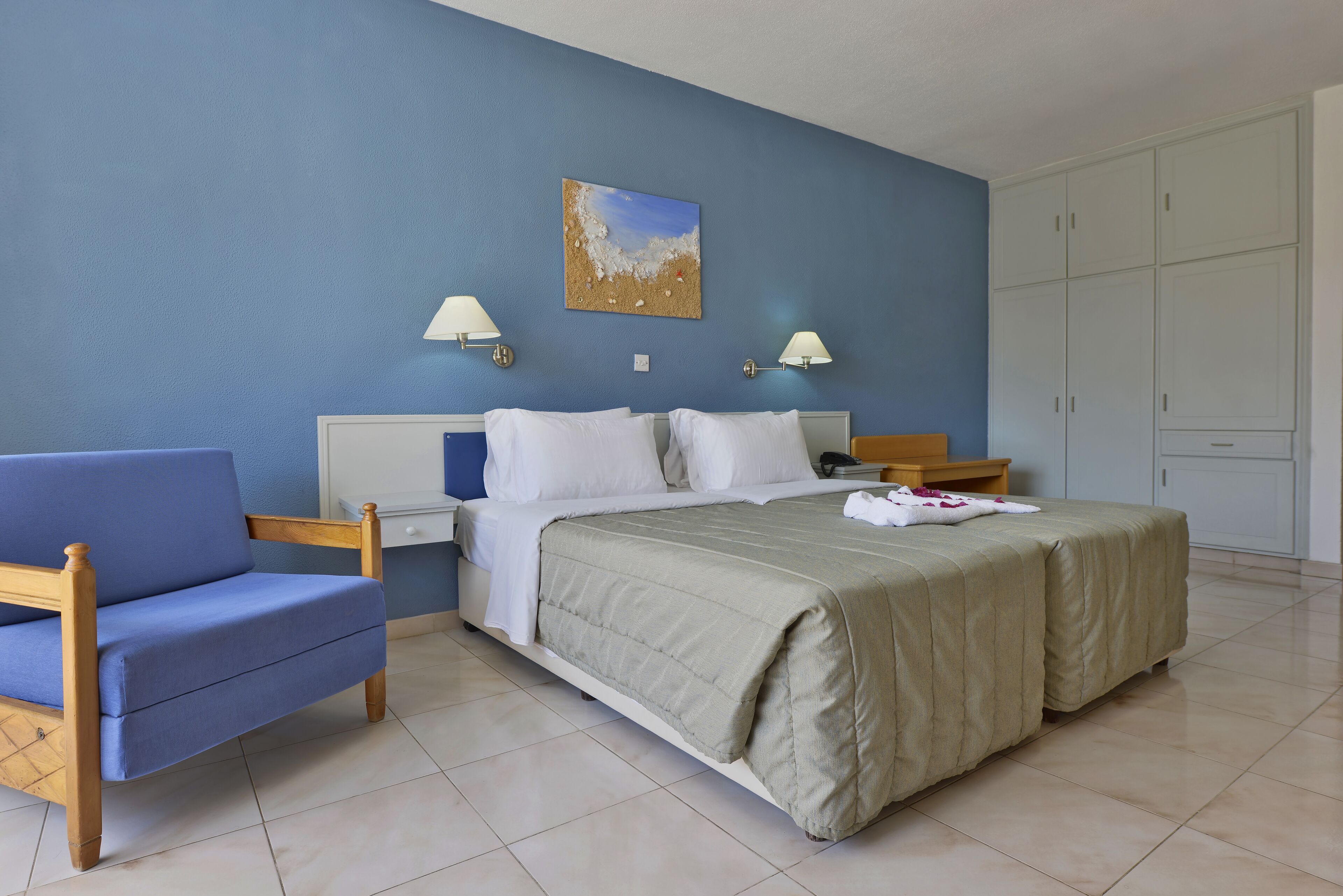 Um quarto com cama, poltrona azul, duas mesas de cabeceira, um criado-mudo e um guarda-roupa.