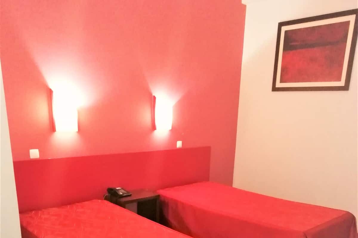 A room with two beds, each covered with a red bedspread. There are two wall-mounted lights above the beds, and a framed picture on the wall to the right.