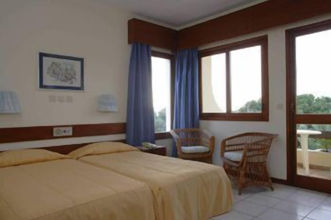 Habitación de hotel con dos camas, una silla, una mesa y un ventanal grande con cortinas.