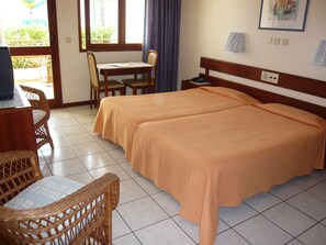 Ein Hotelzimmer mit zwei Betten, einem Schreibtisch mit Stühlen, einem Fenster mit Vorhängen und einem an der Wand hängenden Bild.