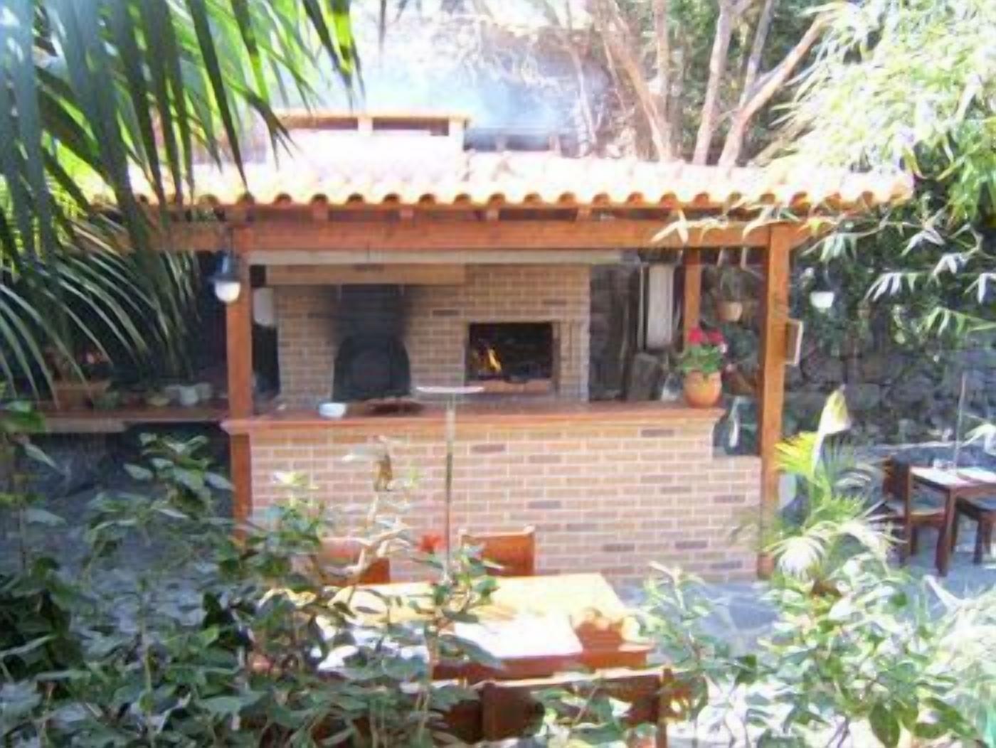 An outdoor brick fireplace with a wooden pergola roof, surrounded by greenery and a table with chairs.
