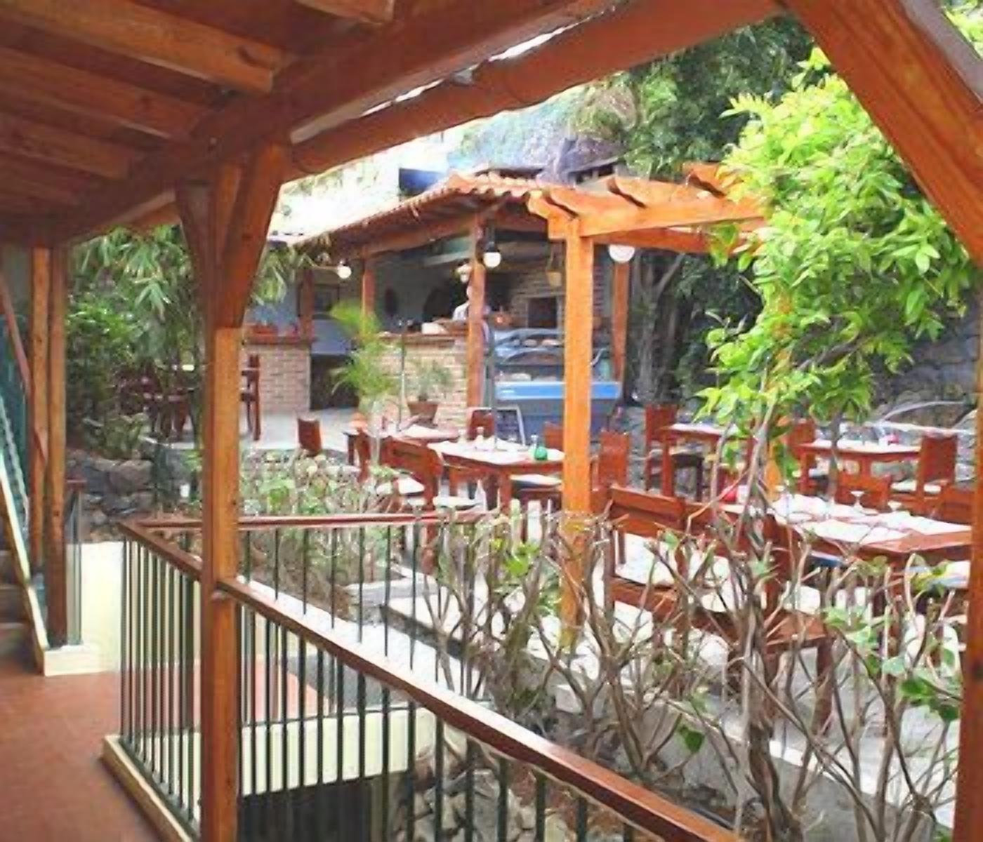 An outdoor patio with wooden tables and chairs, surrounded by plants and a stone wall.